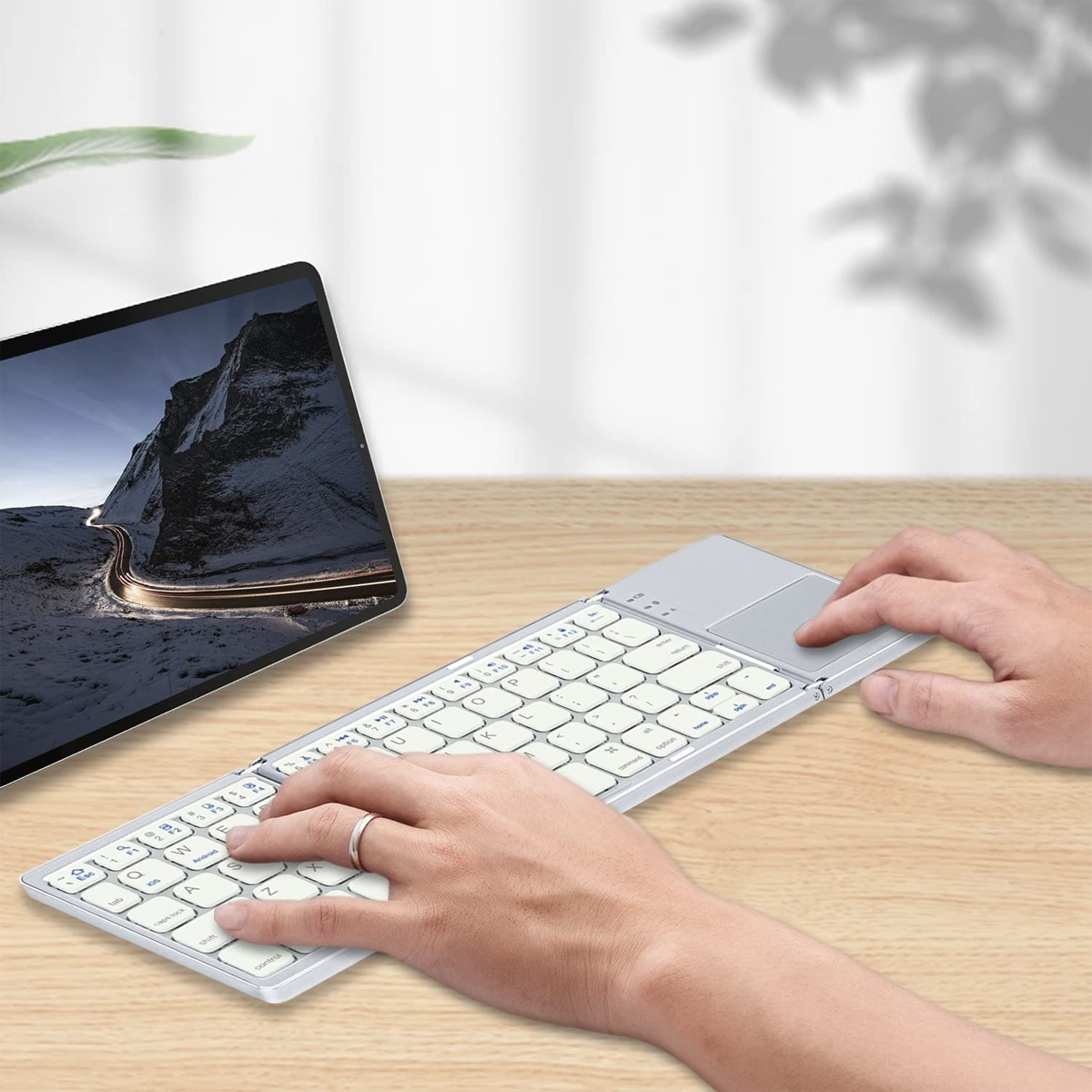 Osoba używająca srebrnej klawiatury z panelem dotykowym. Tablet z obrazem gór w tle.