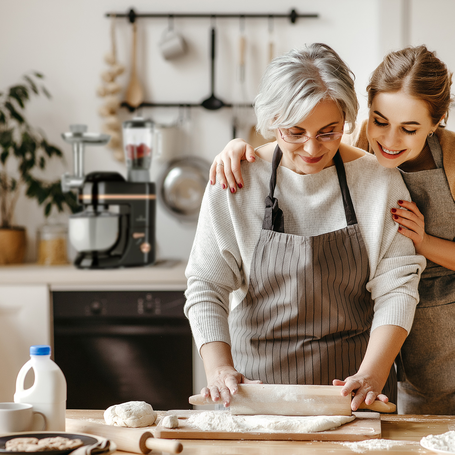 Zwei Frauen backen. Eine rollt Teig, die andere umarmt sie. Küche mit Küchenmaschine im Hintergrund.