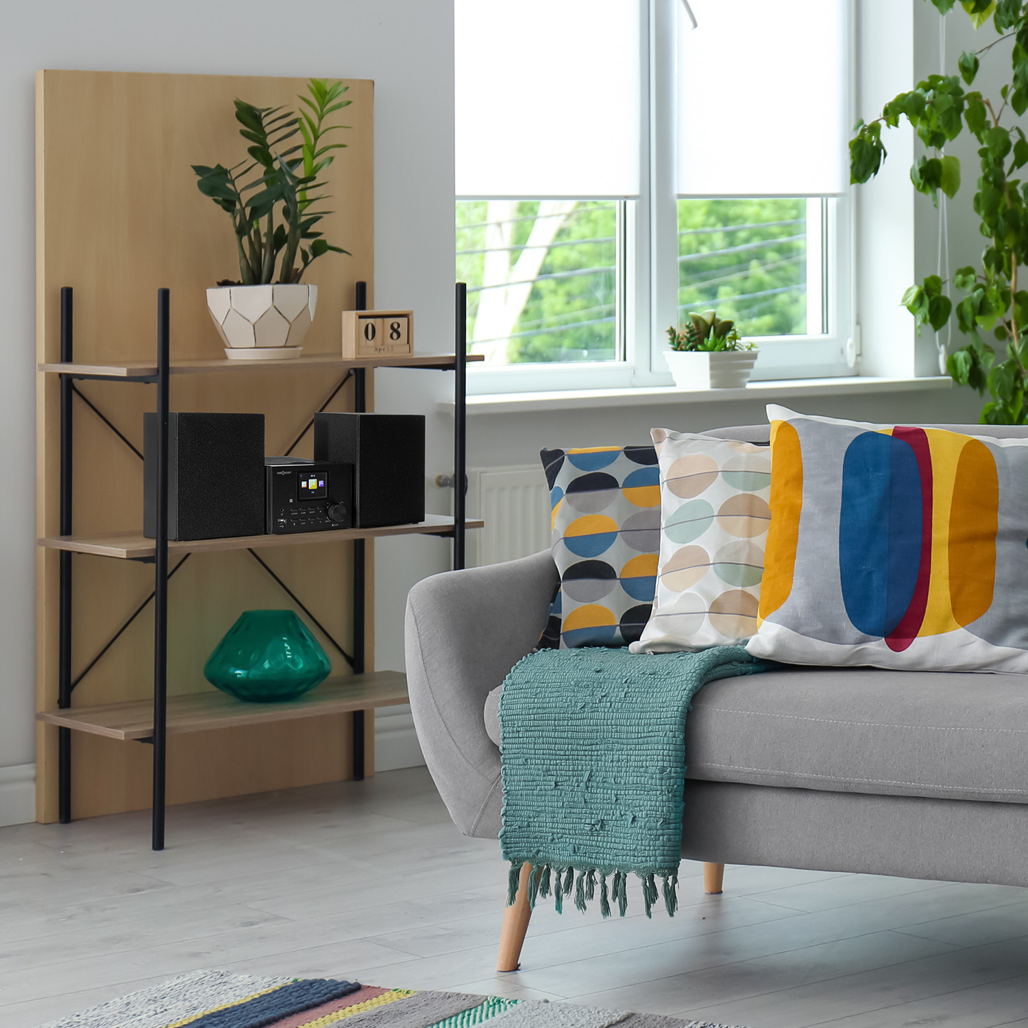 Ein Wohnzimmer zeigt ein Sofa mit Kissen, ein Bücherregal mit Lautsprechern und ein Fenster. Die Szene wirkt natürlich und gemütlich.