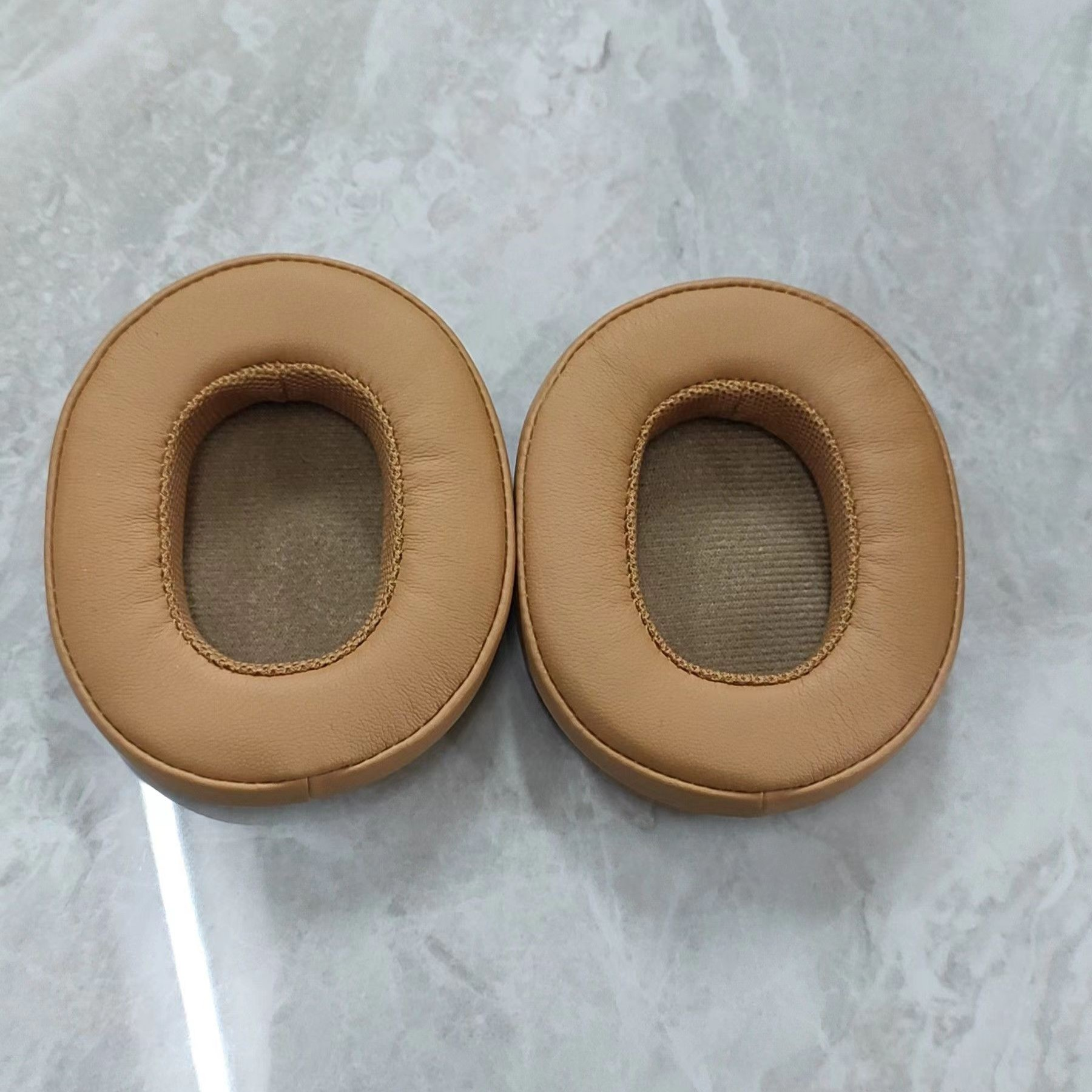 Twee beige, ovaalvormige oorkussens voor een hoofdtelefoon op een lichtgrijze ondergrond.