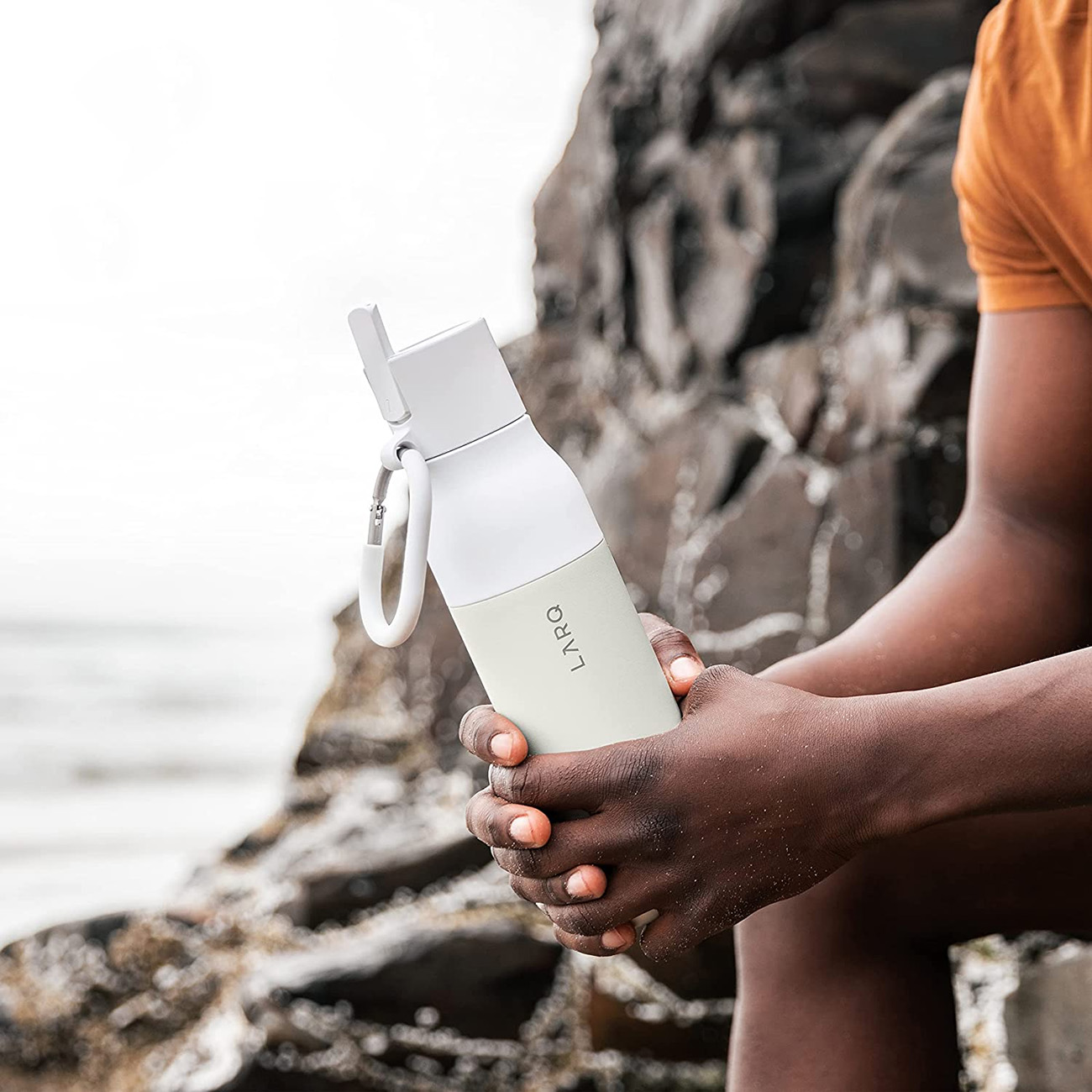 Hände einer Person halten eine weiß-cremefarbene Wasserflasche an einem Strand mit Felsen und Meer im Hintergrund.