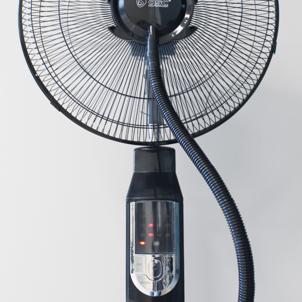 Un ventilador de pie negro contra una pared blanca. El ventilador tiene una pantalla digital y una manguera curva.