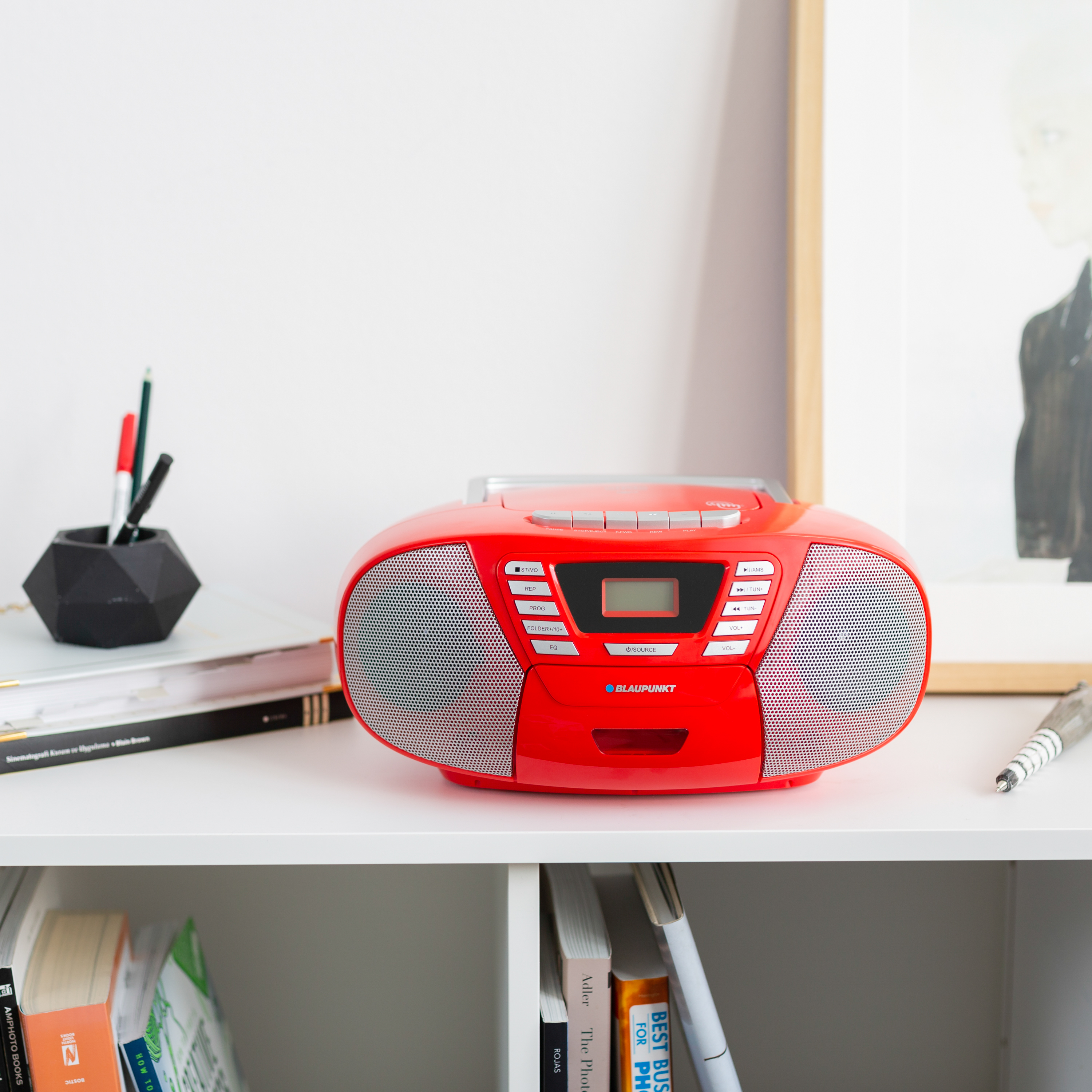 Roter Blaupunkt-Boombox auf weißem Regal. Bücher, Stifte und ein Bilderrahmen sind ebenfalls sichtbar. Weißer Wandhintergrund.