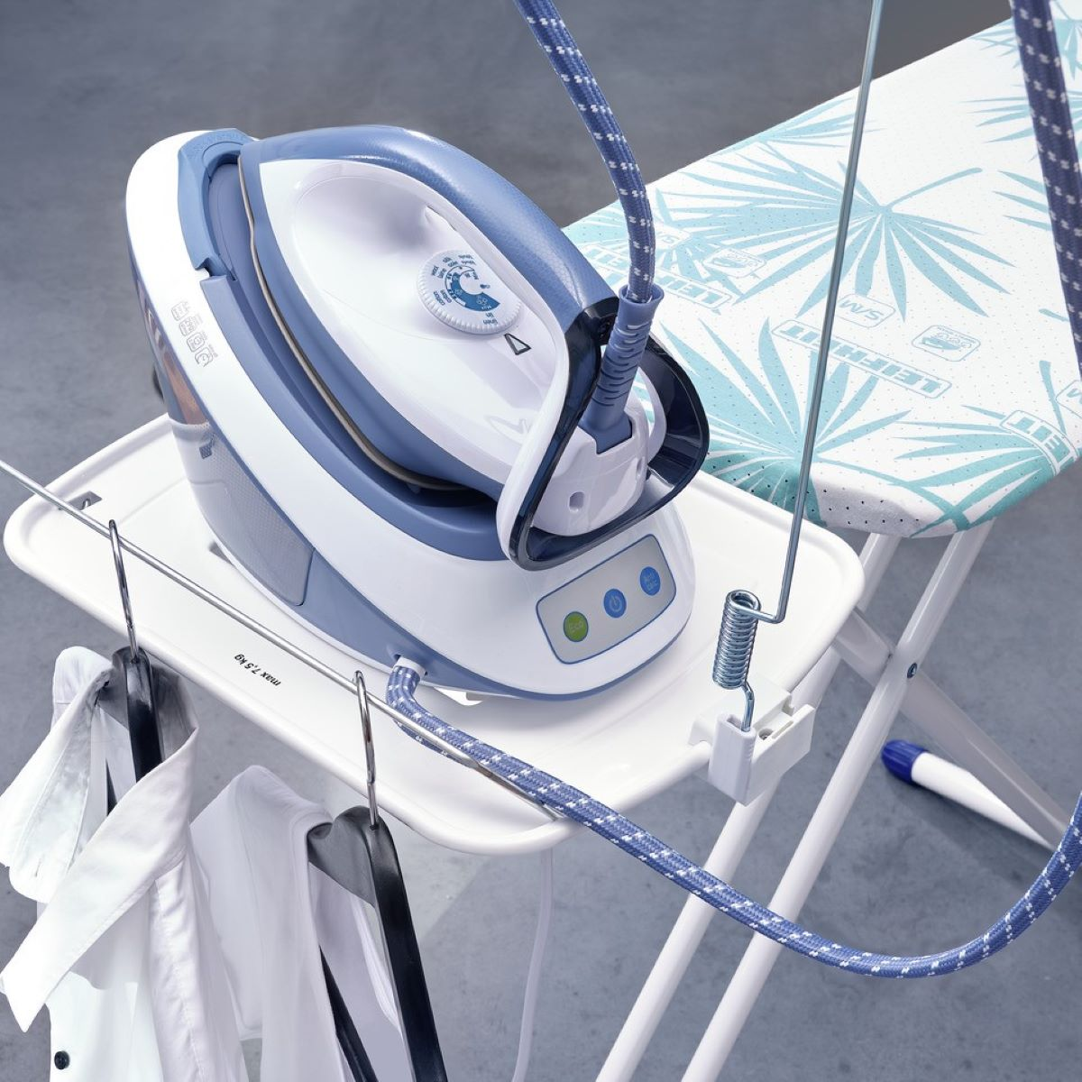 Una plancha azul y blanca sobre una tabla de planchar blanca. Perchas y ropa en primer plano.