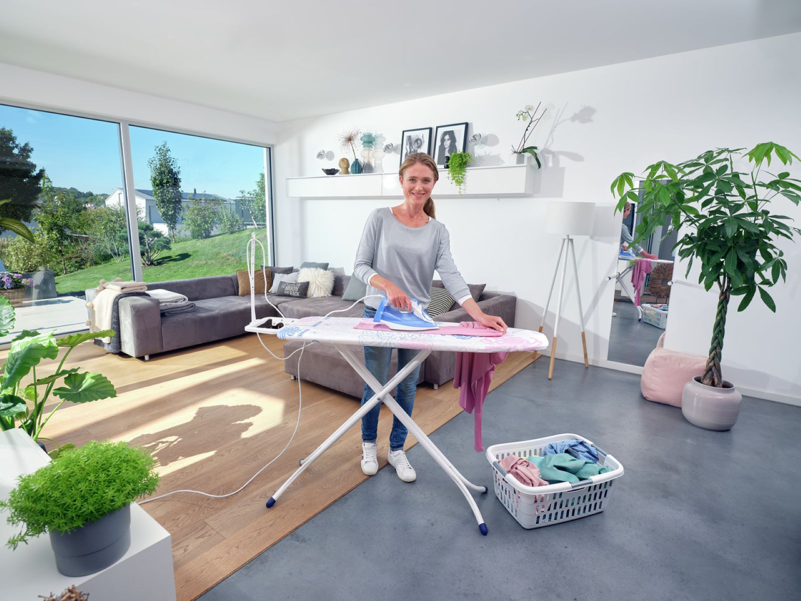 Mujer planchando ropa en una tabla de planchar blanca. Cesta de ropa, plantas y ventana visibles.