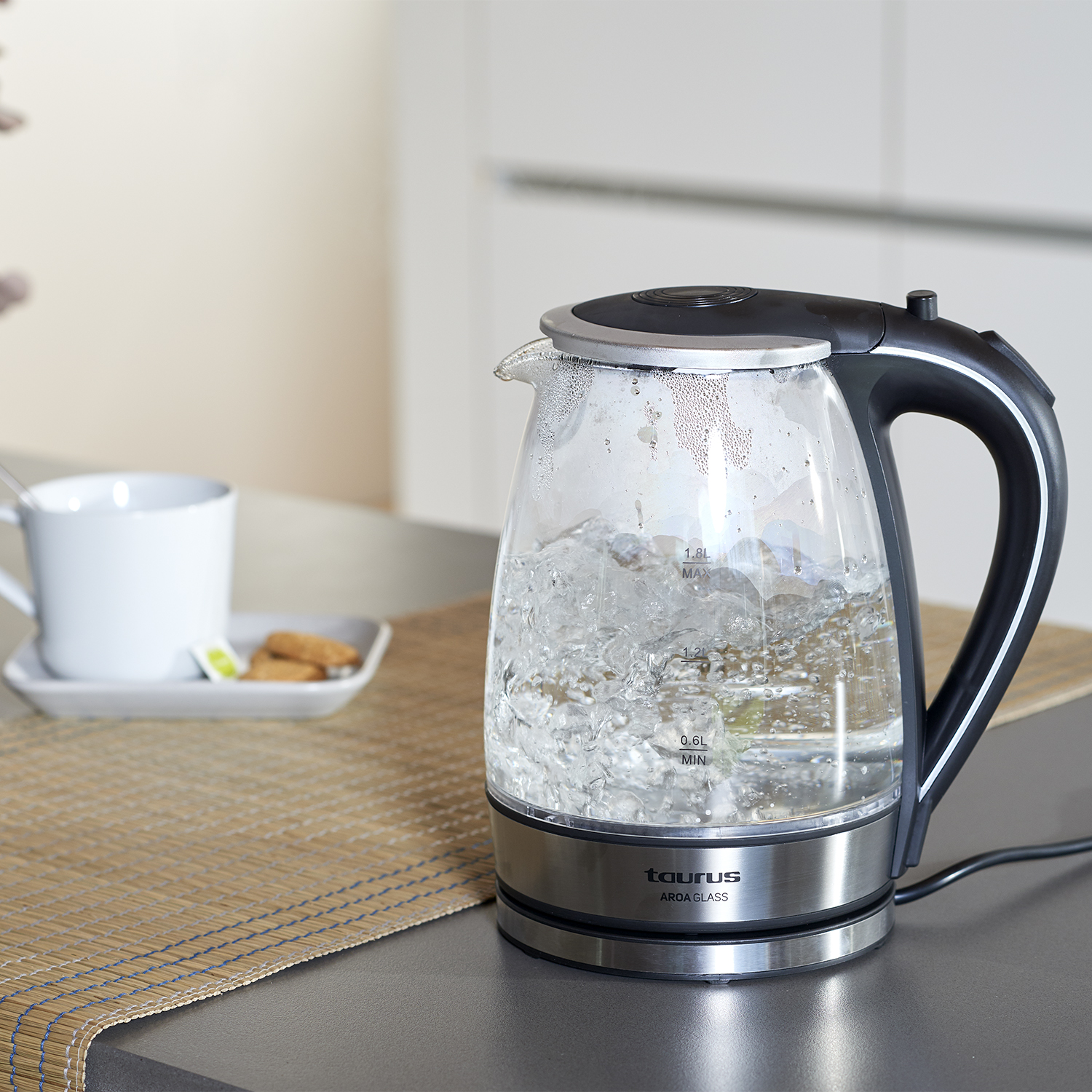 Una tetera eléctrica de vidrio con agua hirviendo, sobre una encimera con una taza y galletas.