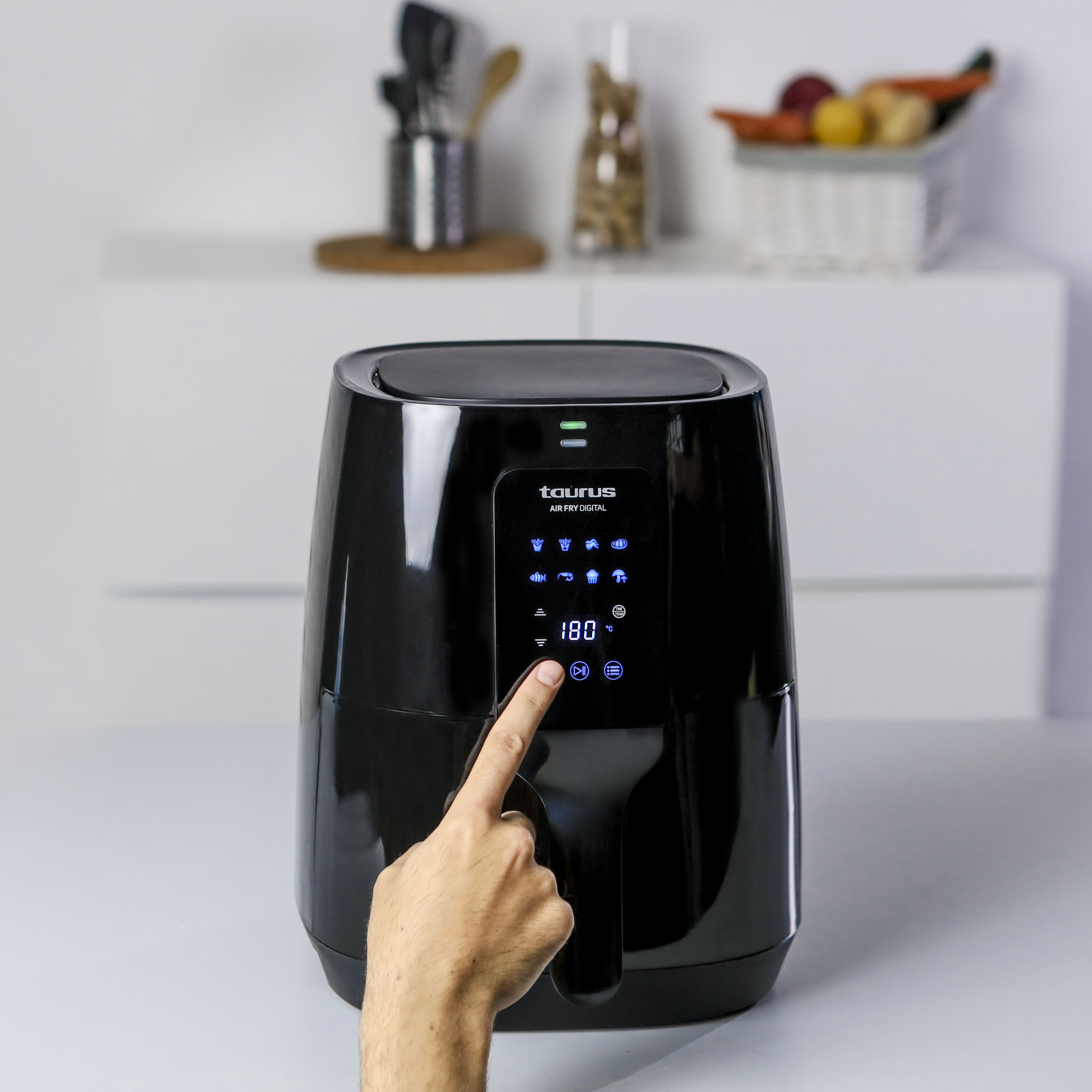 Freidora de aire Taurus negra con pantalla digital, una mano presiona un botón, sobre superficie clara.