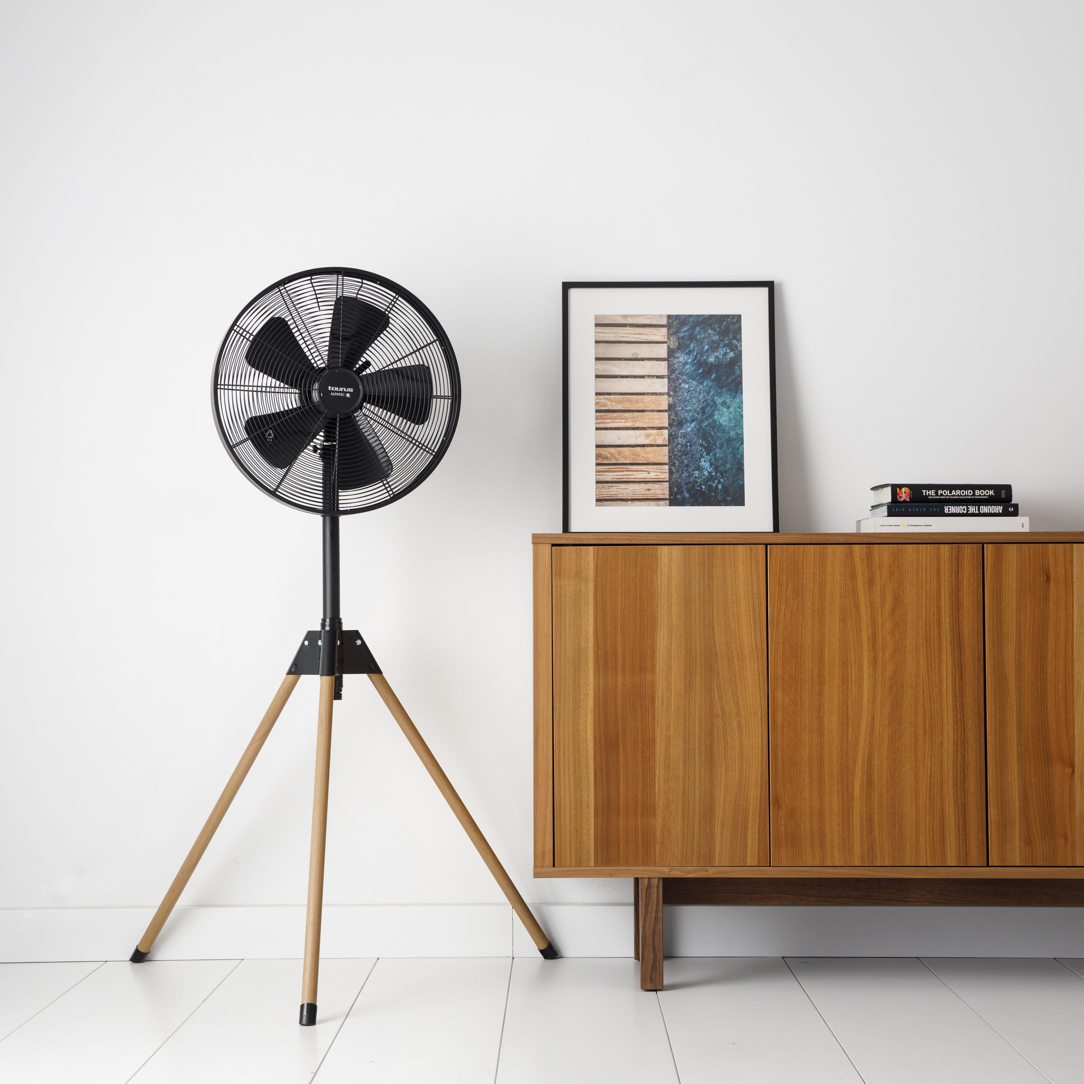 Un ventilador negro en un trípode, junto a un armario y un cuadro en una pared blanca.