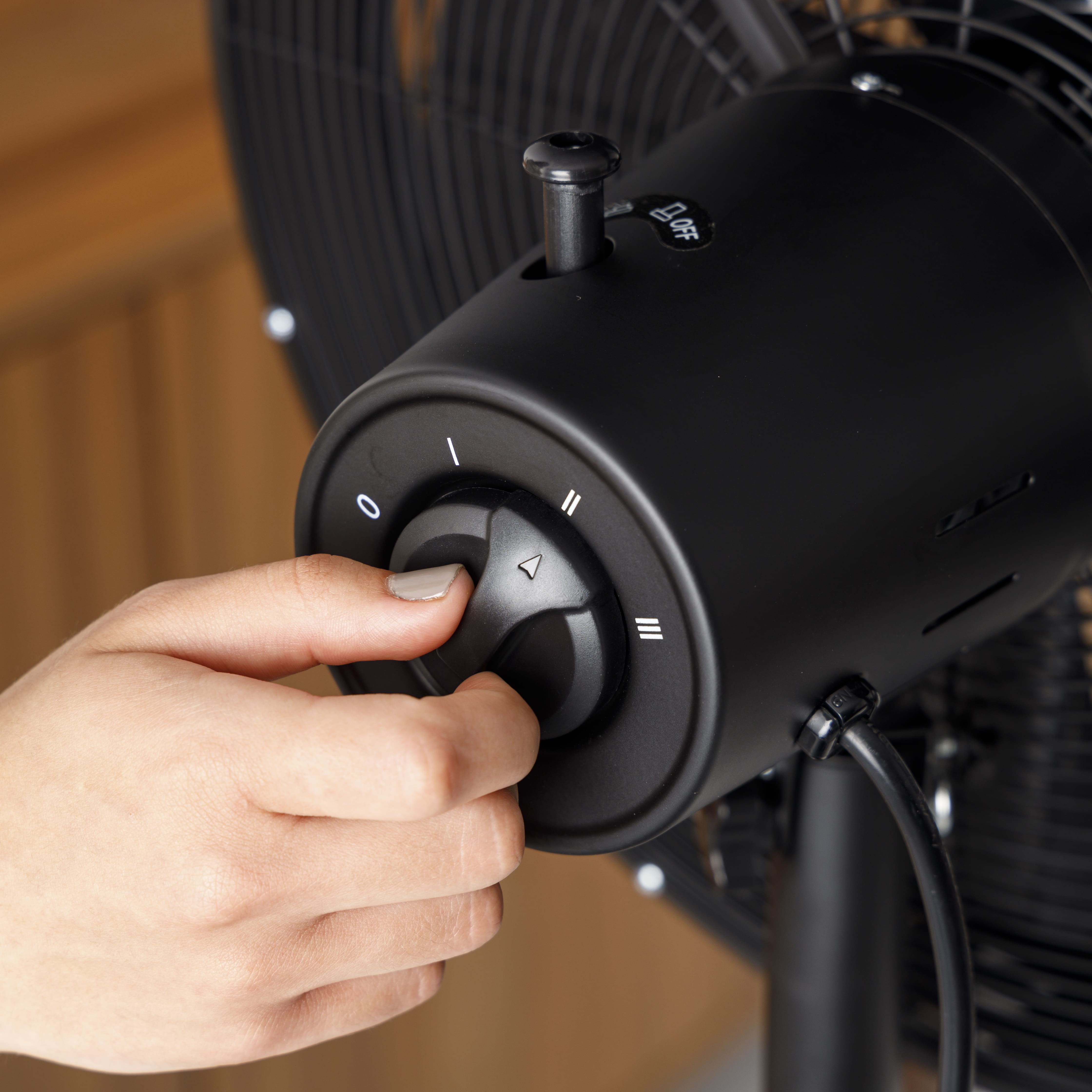 Mano girando el dial de velocidad en un ventilador negro. El dial tiene números y configuraciones. El fondo es de madera.