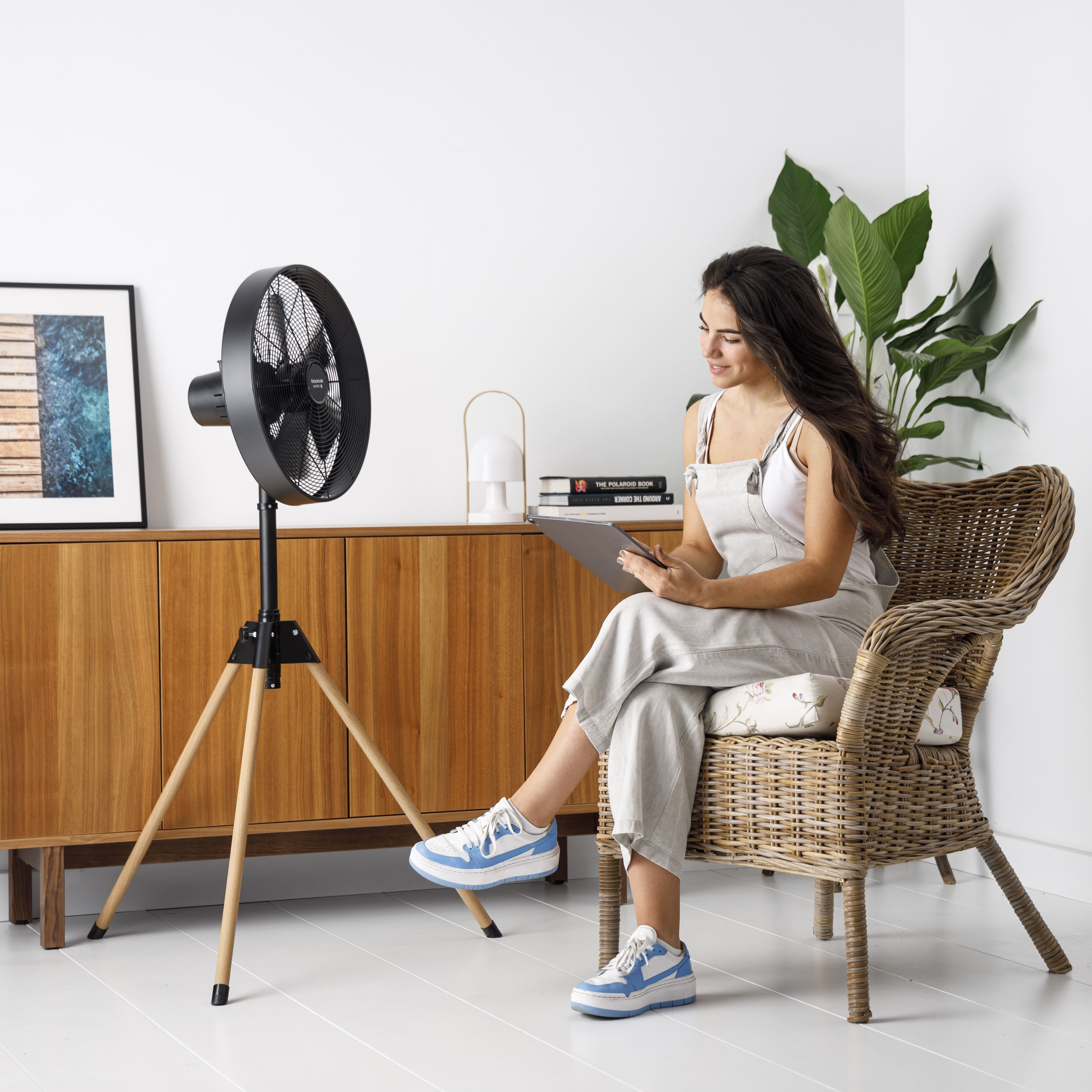 Mujer con tableta sentada junto a ventilador en trípode. Detrás, mueble de madera y planta.