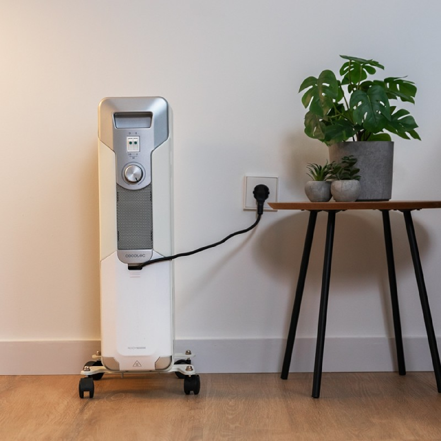 Biały grzejnik elektryczny z roślinami na stole. Jest gniazdko ścienne i drewniana podłoga.