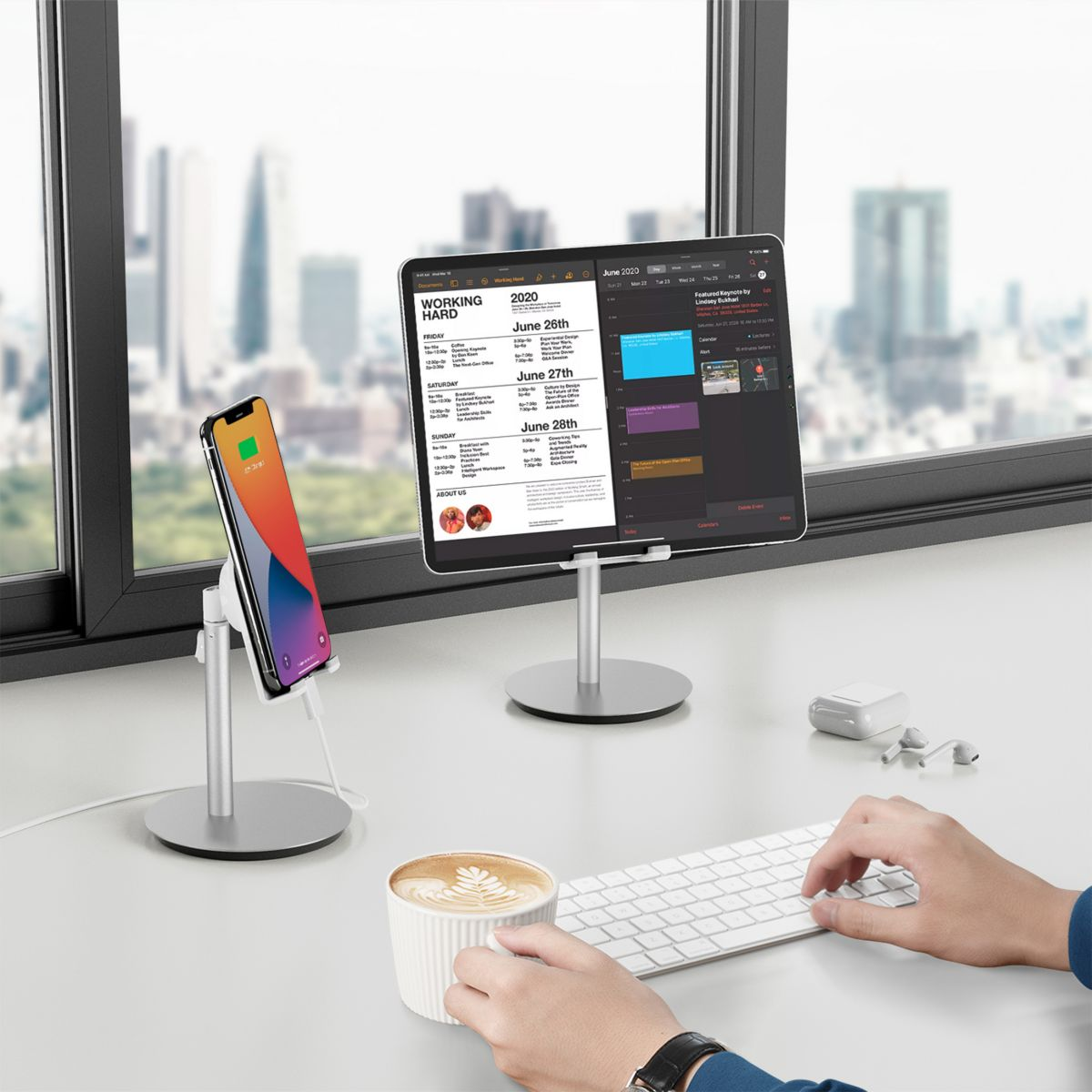 Schreibtisch mit Tablet, Telefon und Kaffee. Tastatur und Hände sichtbar. Stadtblick im Hintergrund.