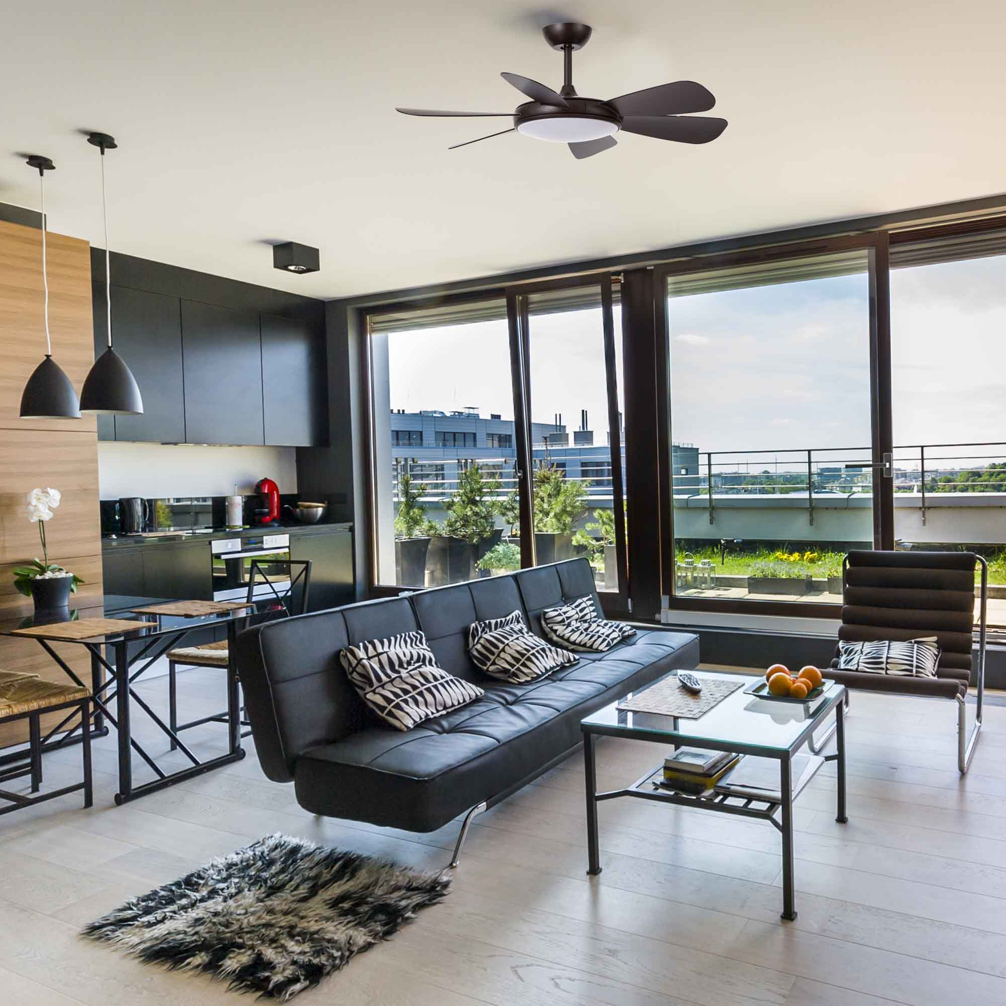 Una sala de estar con ventilador de techo. Hay un sofá, una mesa de cristal y ventanales.