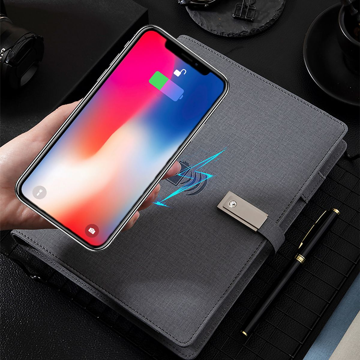 Ein Telefon, das auf einem Pad geladen wird, neben einem Stift und einem Notizbuch, auf einem Schreibtisch.