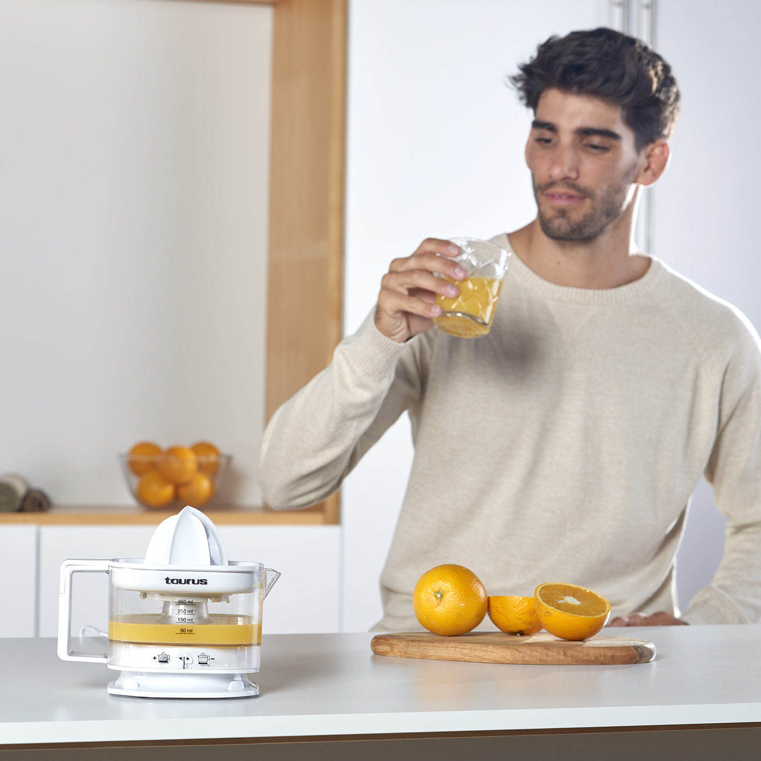 Hombre con vaso de zumo, exprimidor, naranjas en tabla y fondo de cocina.