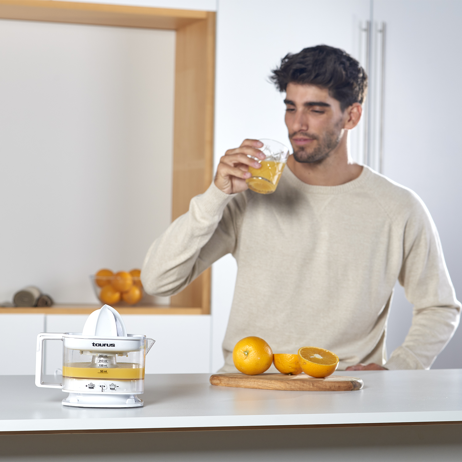 Hombre bebiendo zumo, exprimidor y naranjas en una encimera.