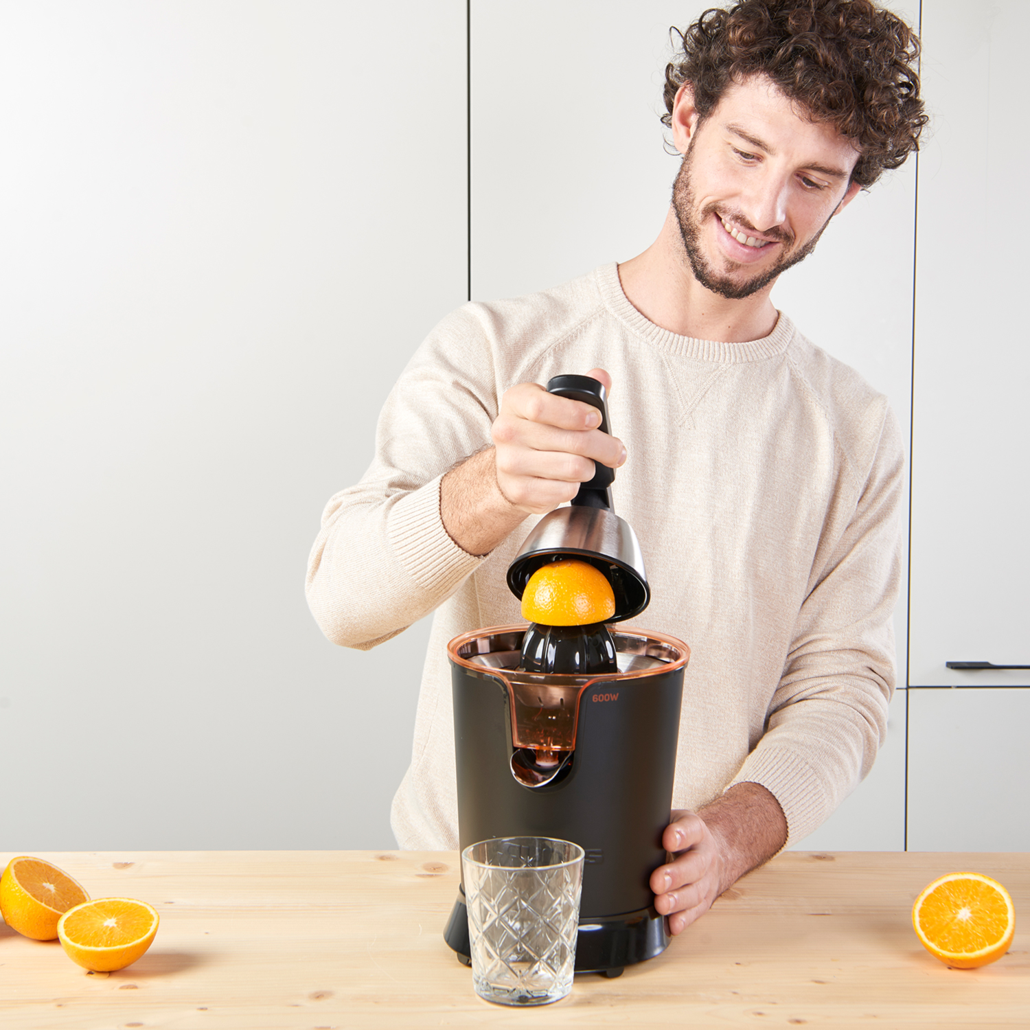 Hombre exprimiendo zumo de naranja con una exprimidora. Hay un vaso y naranjas en una mesa de madera.