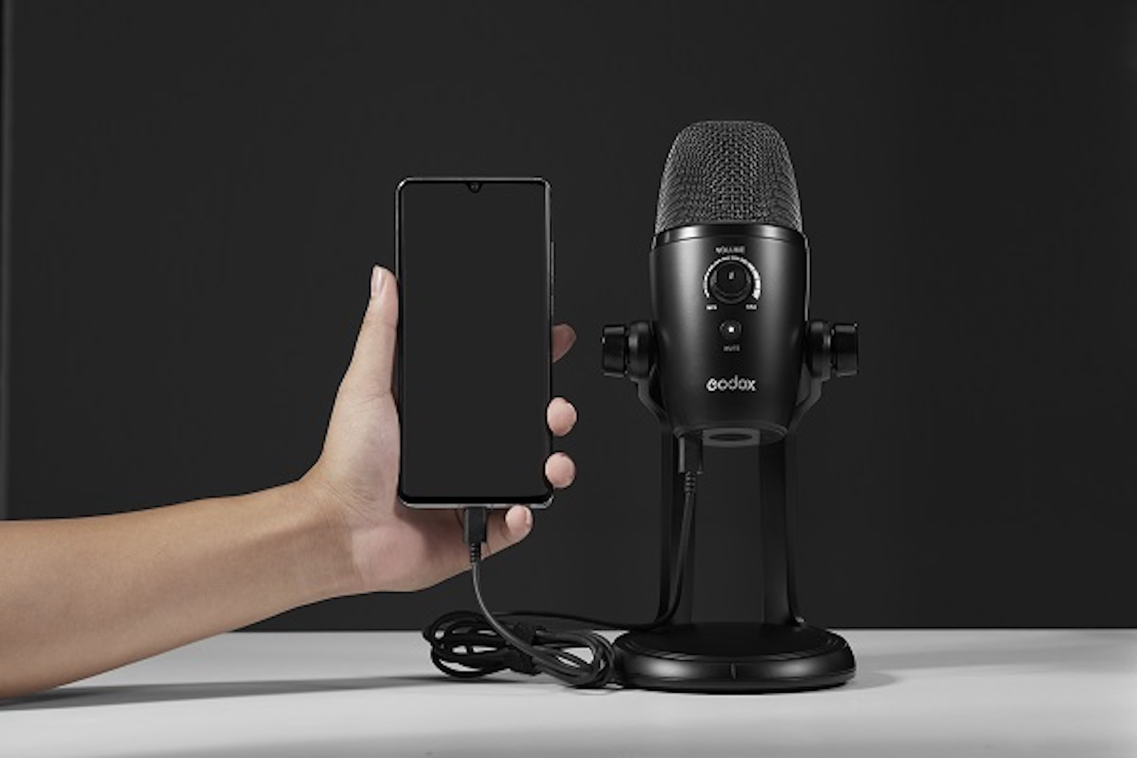 Microphone noir sur pied, connecté à un smartphone tenu en main. Fond noir.