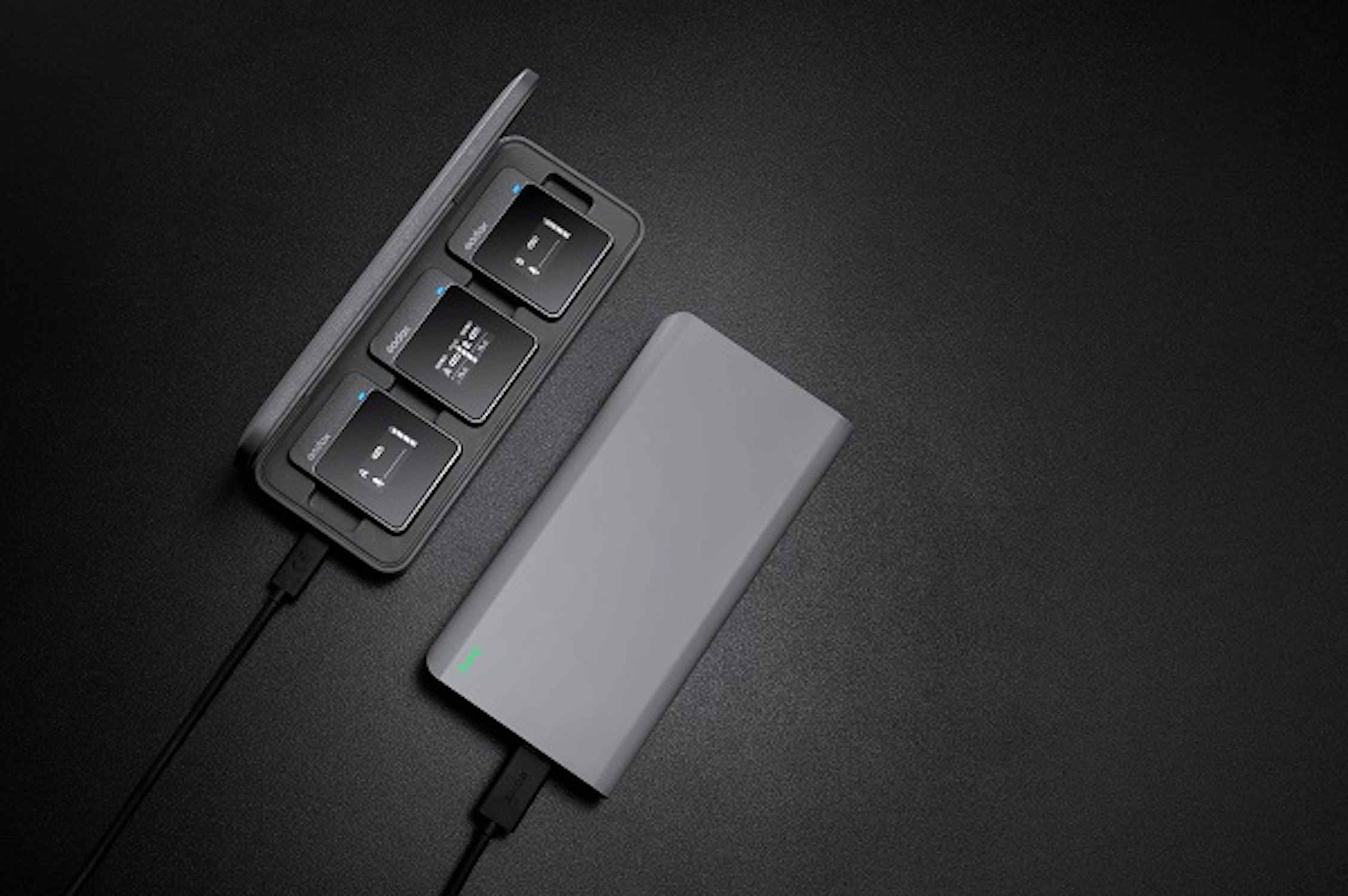 Auf einer schwarzen Oberfläche befinden sich eine Ladestation mit drei Geräten und eine Powerbank. Schwarze Kabel sind angeschlossen.