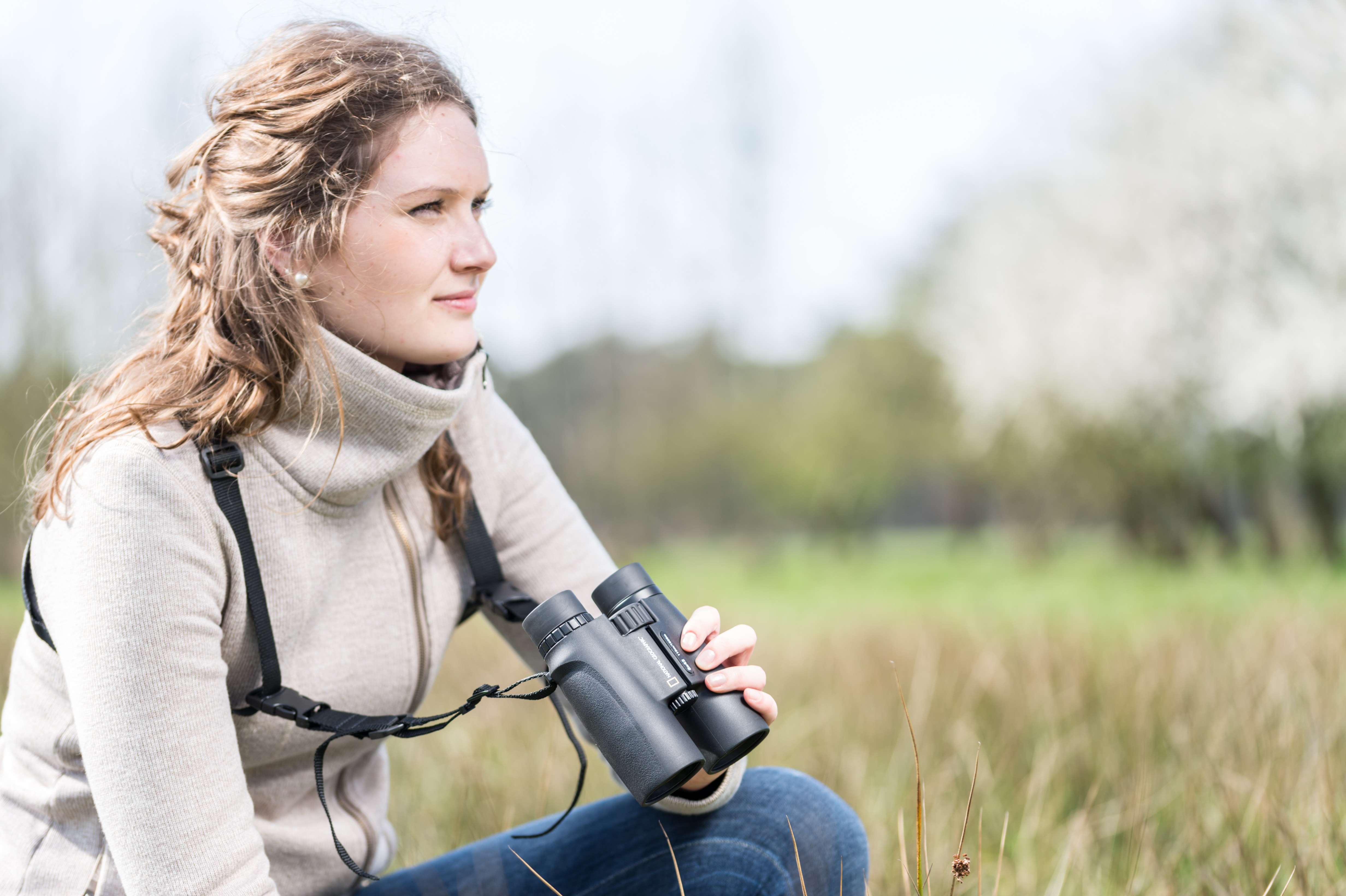 Mujer mirando a través de binoculares al aire libre. Lleva un suéter y vaqueros en un campo.