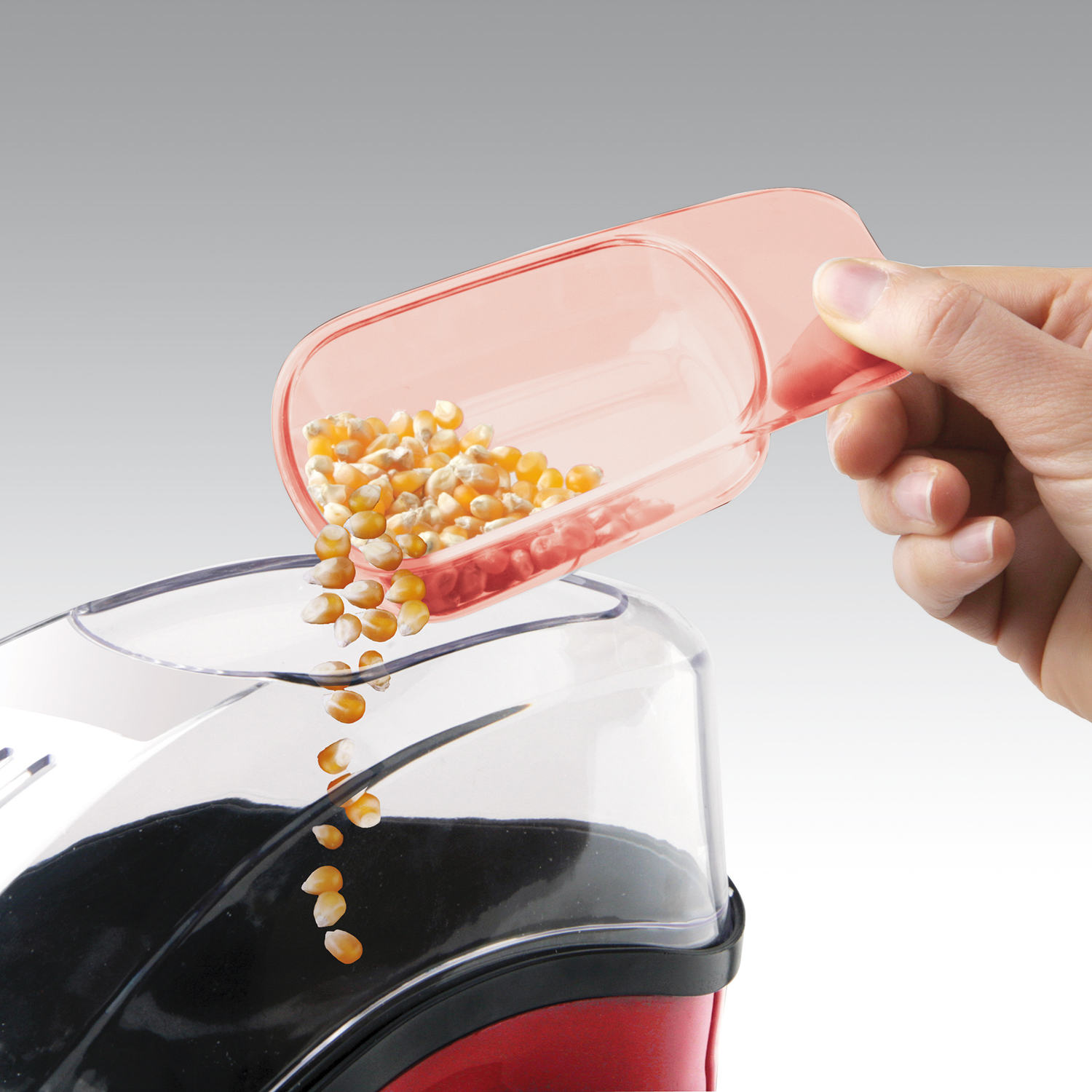 Hand gießt Popcornkerne aus einem rosa Löffel in einen rot-schwarzen Popcornbereiter.