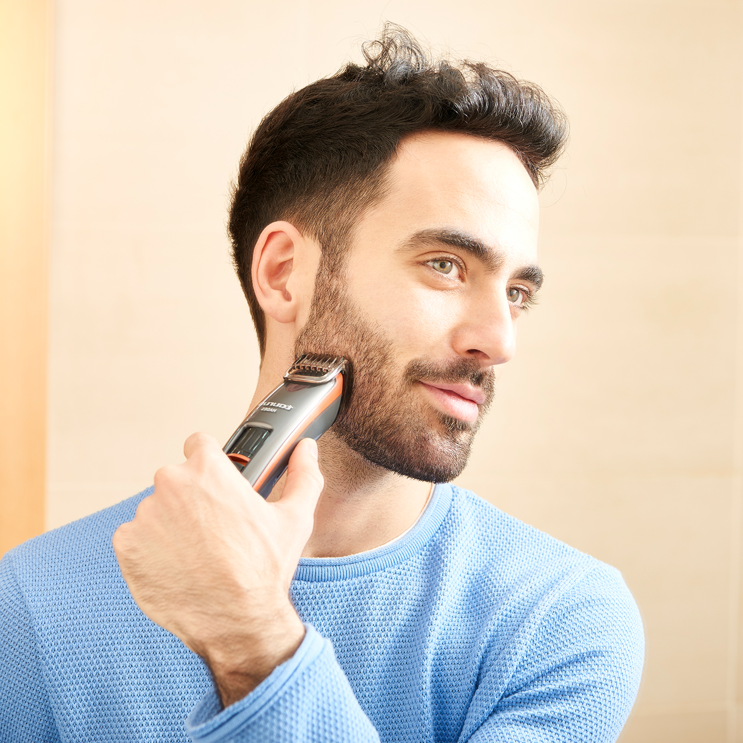 Un hombre se afeita la barba con una recortadora eléctrica. Lleva un suéter azul y se mira al espejo.