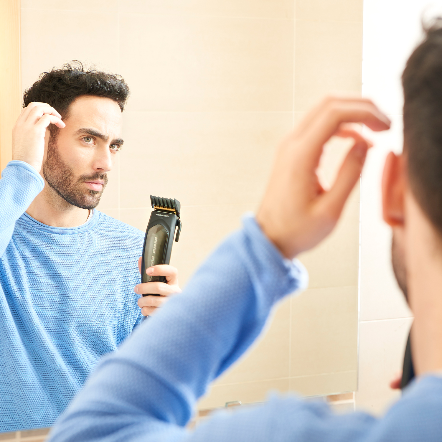 Uomo che si guarda allo specchio, con in mano un tagliacapelli nero. Parete beige e bagno.