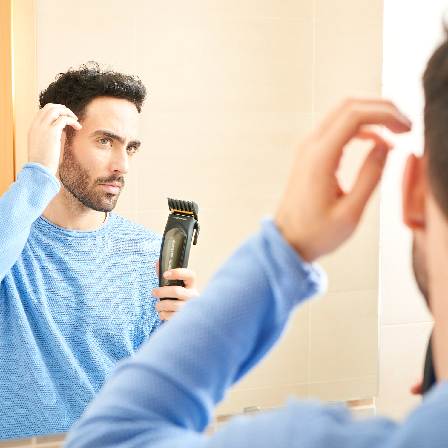 Un hombre mirándose en un espejo, sosteniendo una cortadora de pelo negra y tocándose el pelo, en un baño.