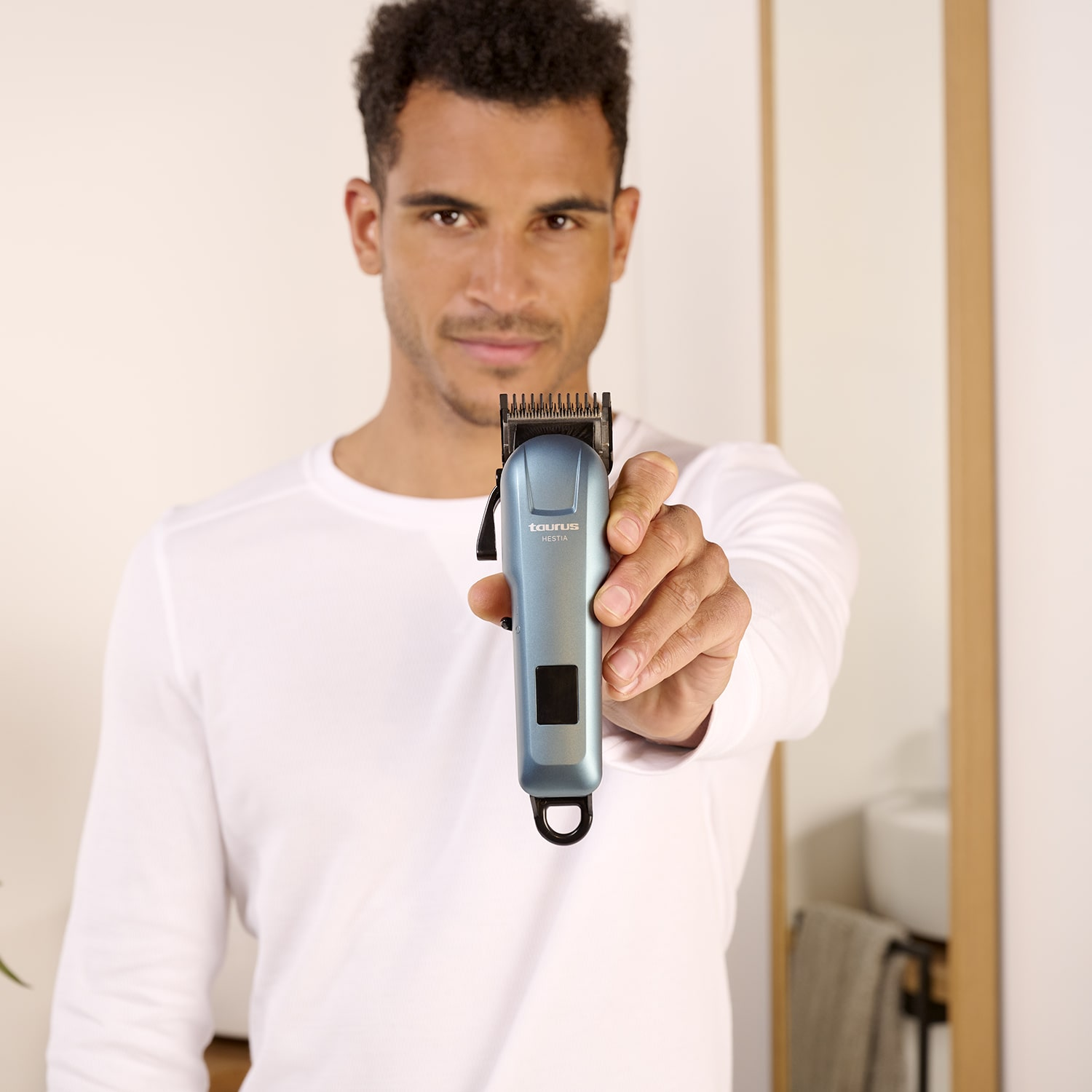 Hombre sostiene una cortadora de cabello azul. Detrás, una pared blanca es visible.