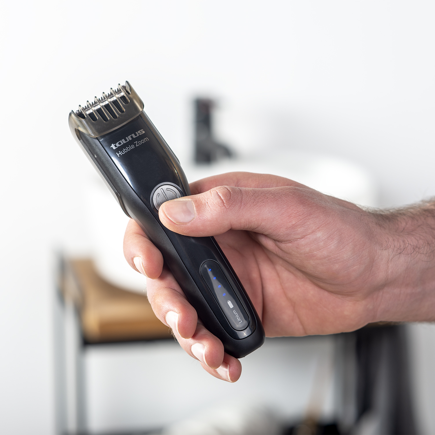 Recortador de barba negro en la mano. El fondo es un lavabo blanco y un banco de madera.
