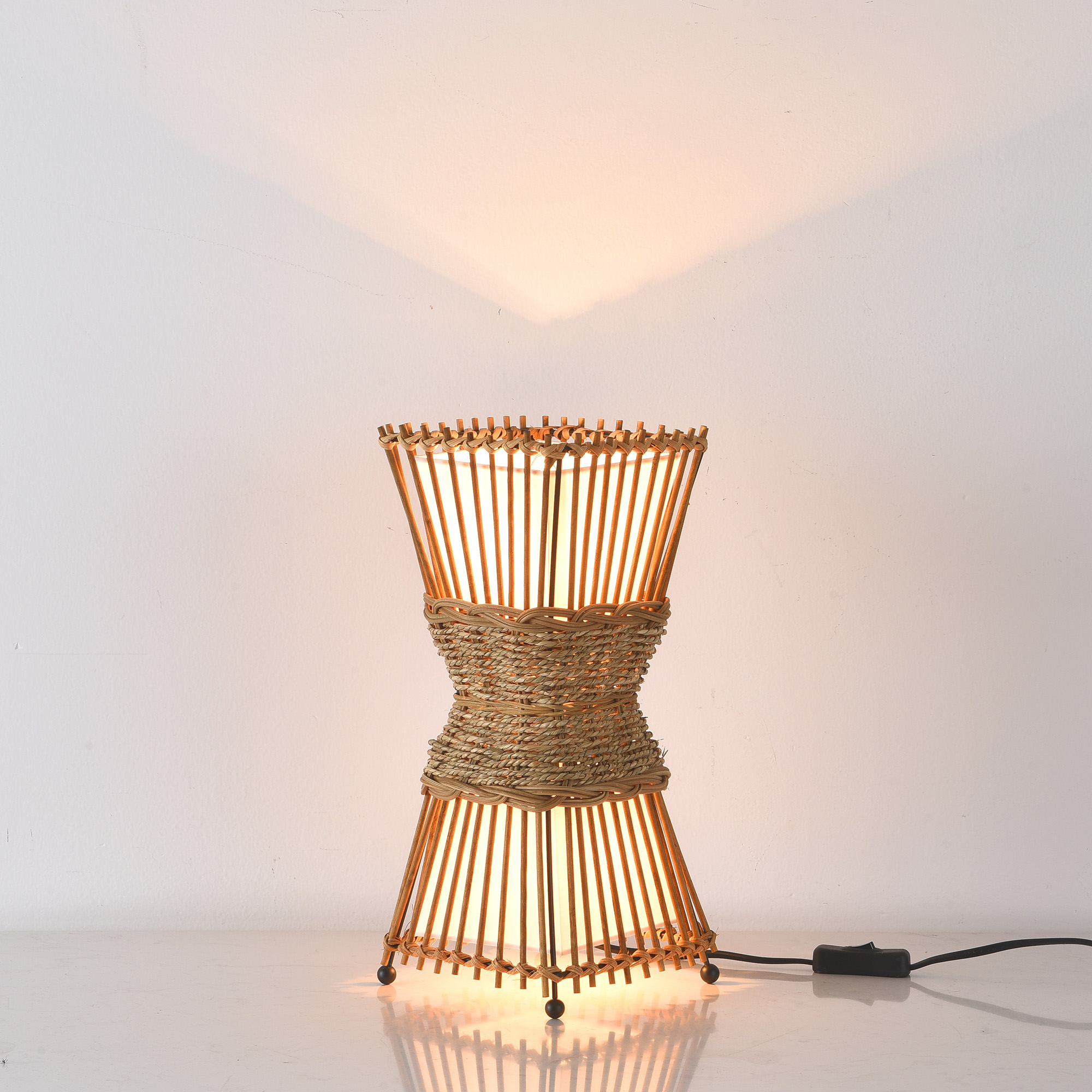Eine dekorative Lampe aus geflochtenem Holz steht auf einer weißen Oberfläche. Das Licht scheint nach oben.