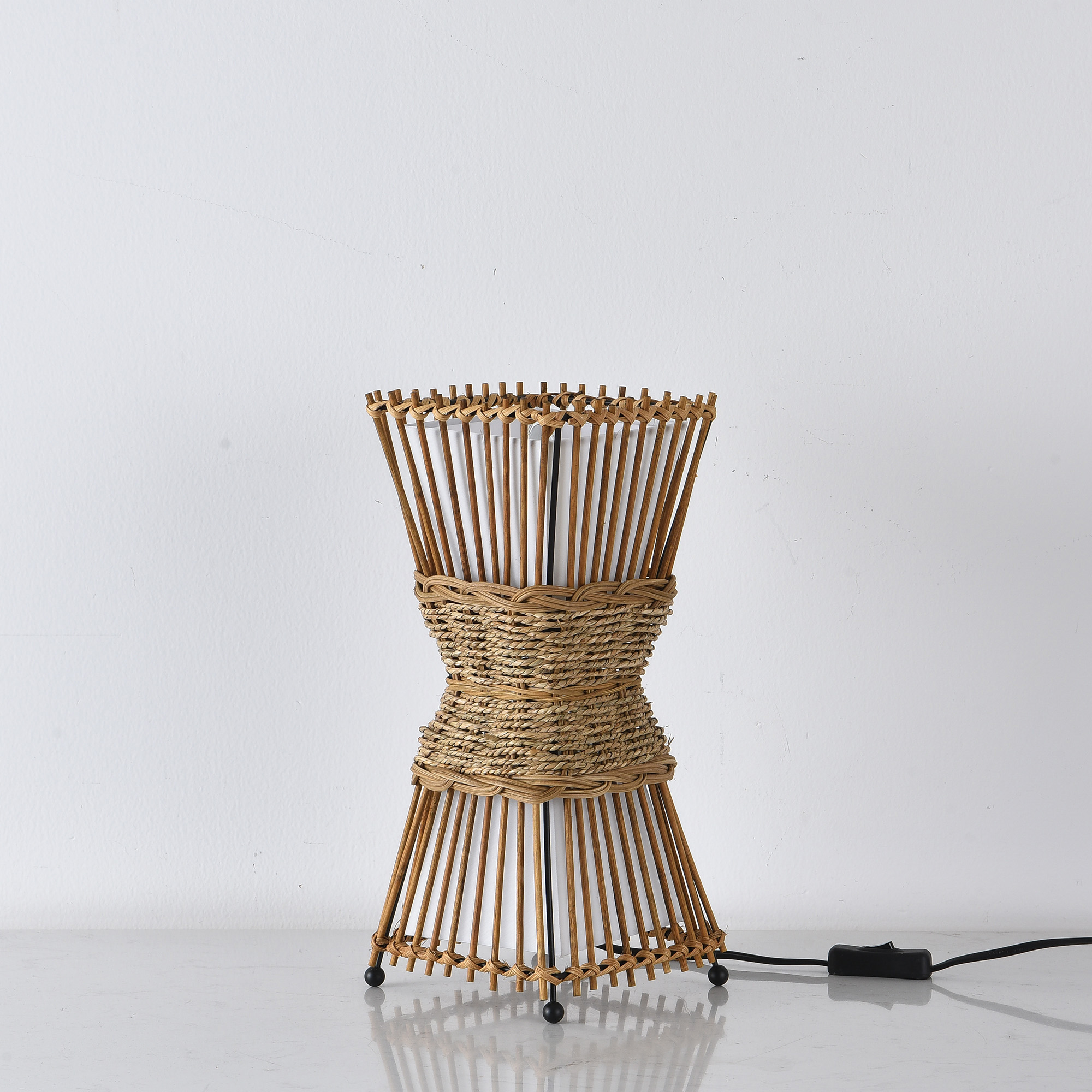 Eine Wicker-Lampe in einer Sanduhrform auf einer weißen Oberfläche und einem weißen Hintergrund.