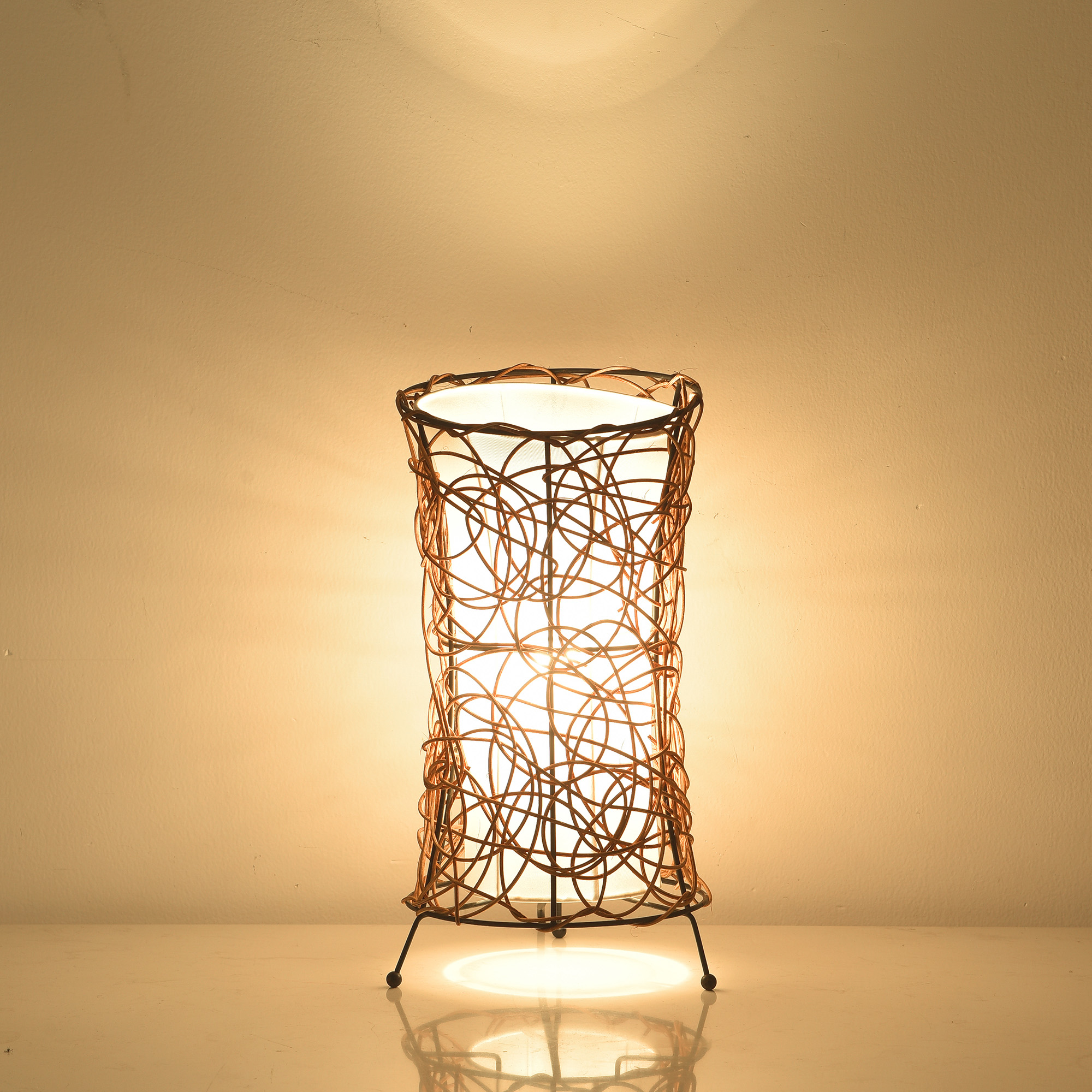Eine zylindrische Tischlampe mit gewebtem Design, von innen beleuchtet. Beige Wand.