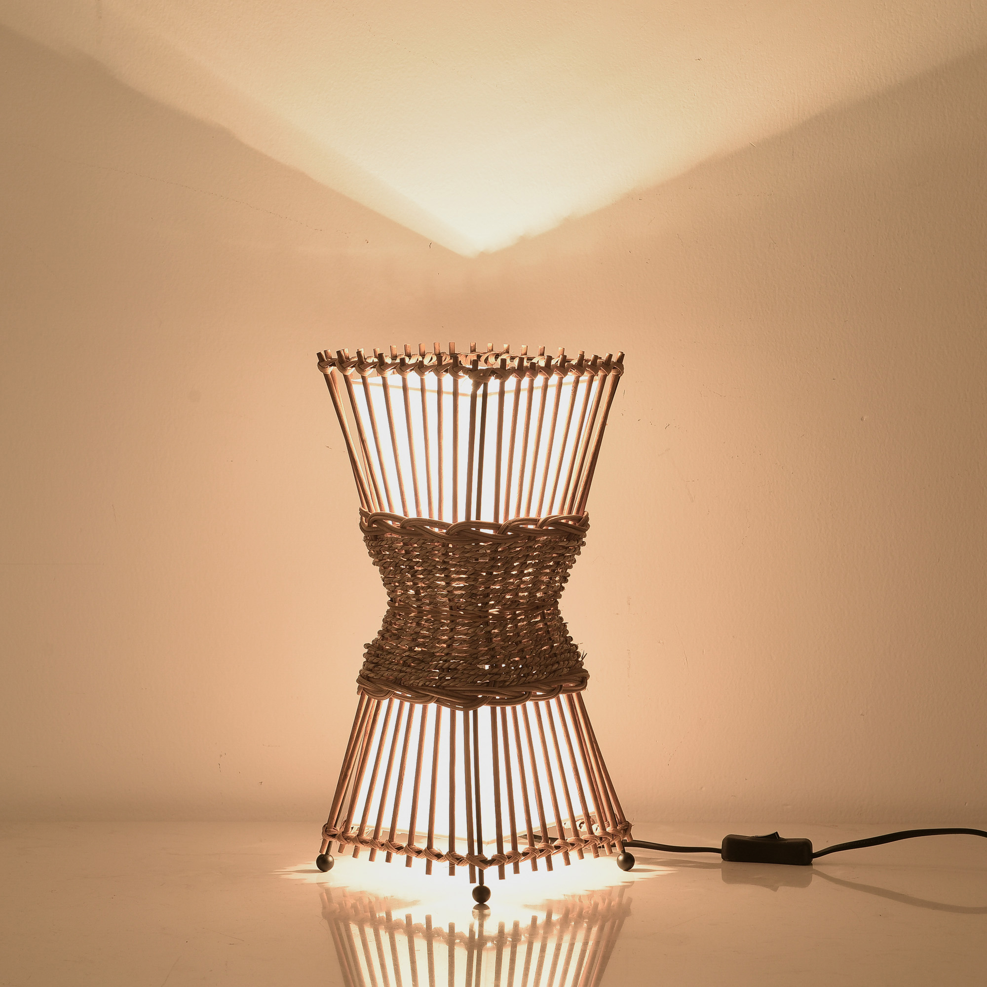 Eine gewebte Lampe, braun und weiß. Sie hat eine Sanduhrform. Licht kommt aus der Lampe.