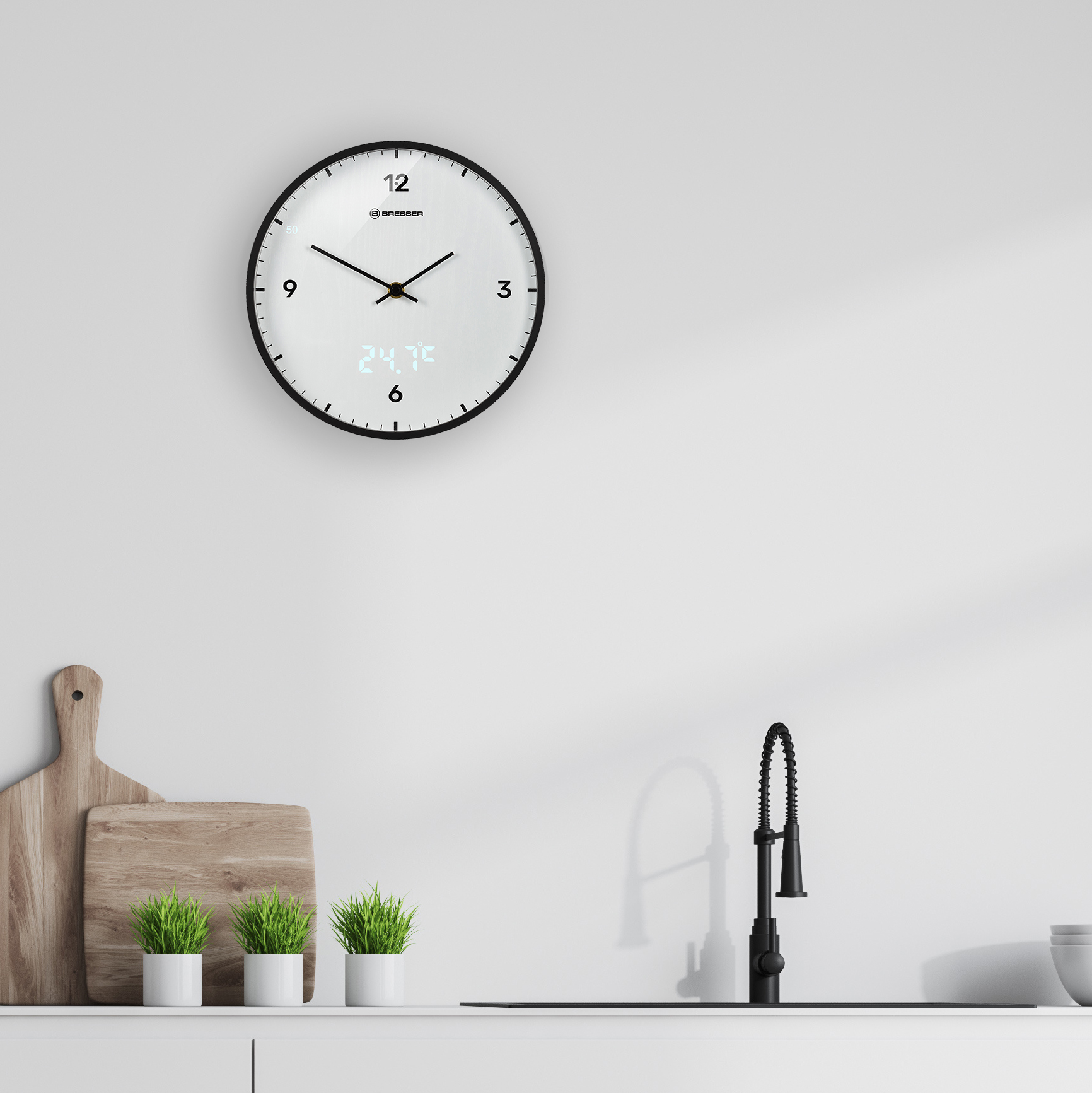 Reloj redondo en una pared blanca, con un grifo negro y dos pequeñas plantas debajo.