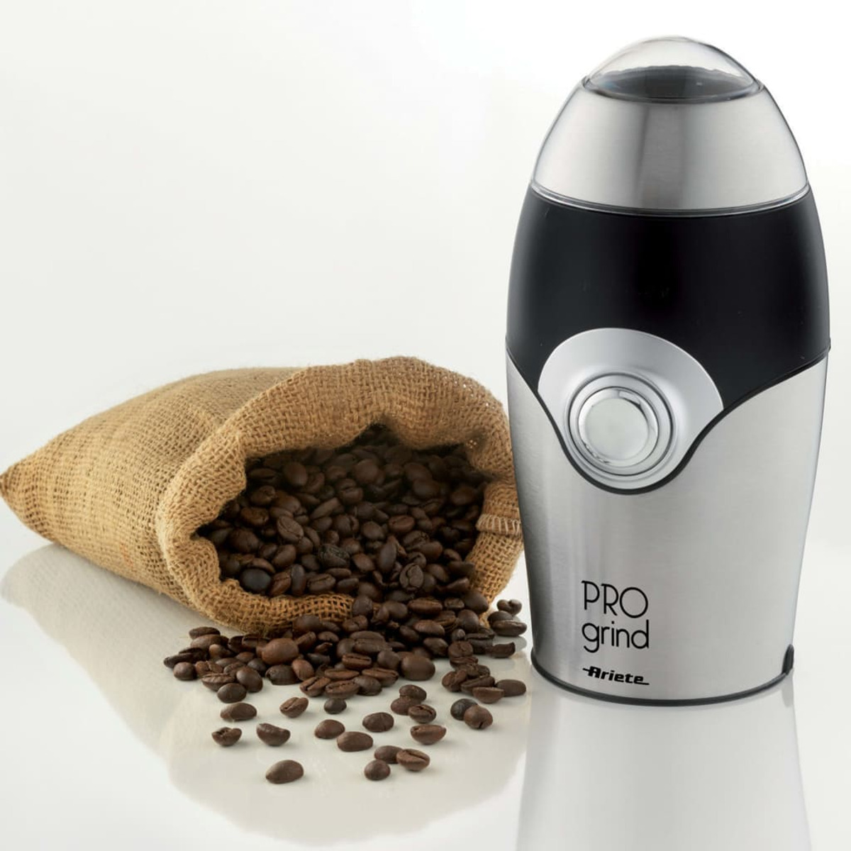 Kaffeebohnen aus einem Jutesack neben einer silber-schwarzen Kaffeemühle. Wort 'PRO GRIND' sichtbar.