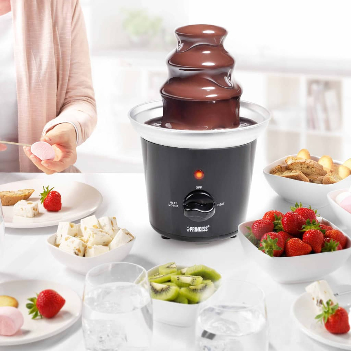 Una fuente de chocolate con frutas y dulces para mojar. Una mujer sostiene una malvavisco.