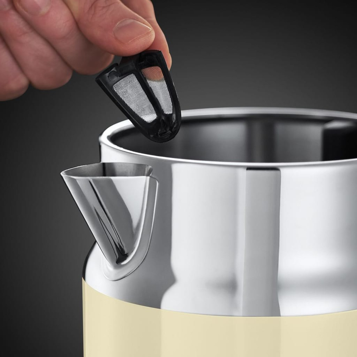 Hand, die einen schwarzen Filter aus einem cremefarbenen Wasserkocher mit silbernem Ausguss entfernt, auf dunklem Hintergrund.