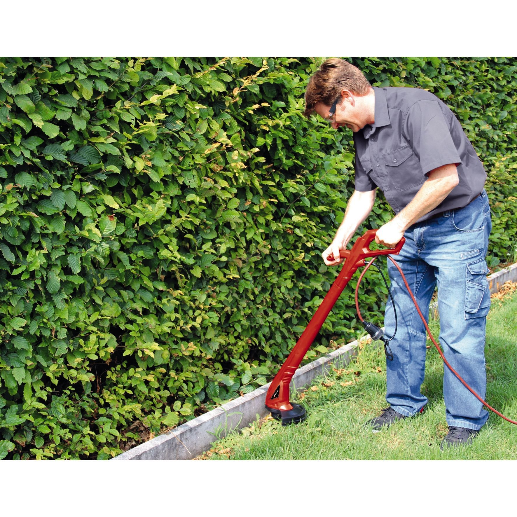 Mann trimmt Gras mit rotem Trimmer neben einer grünen Hecke.
