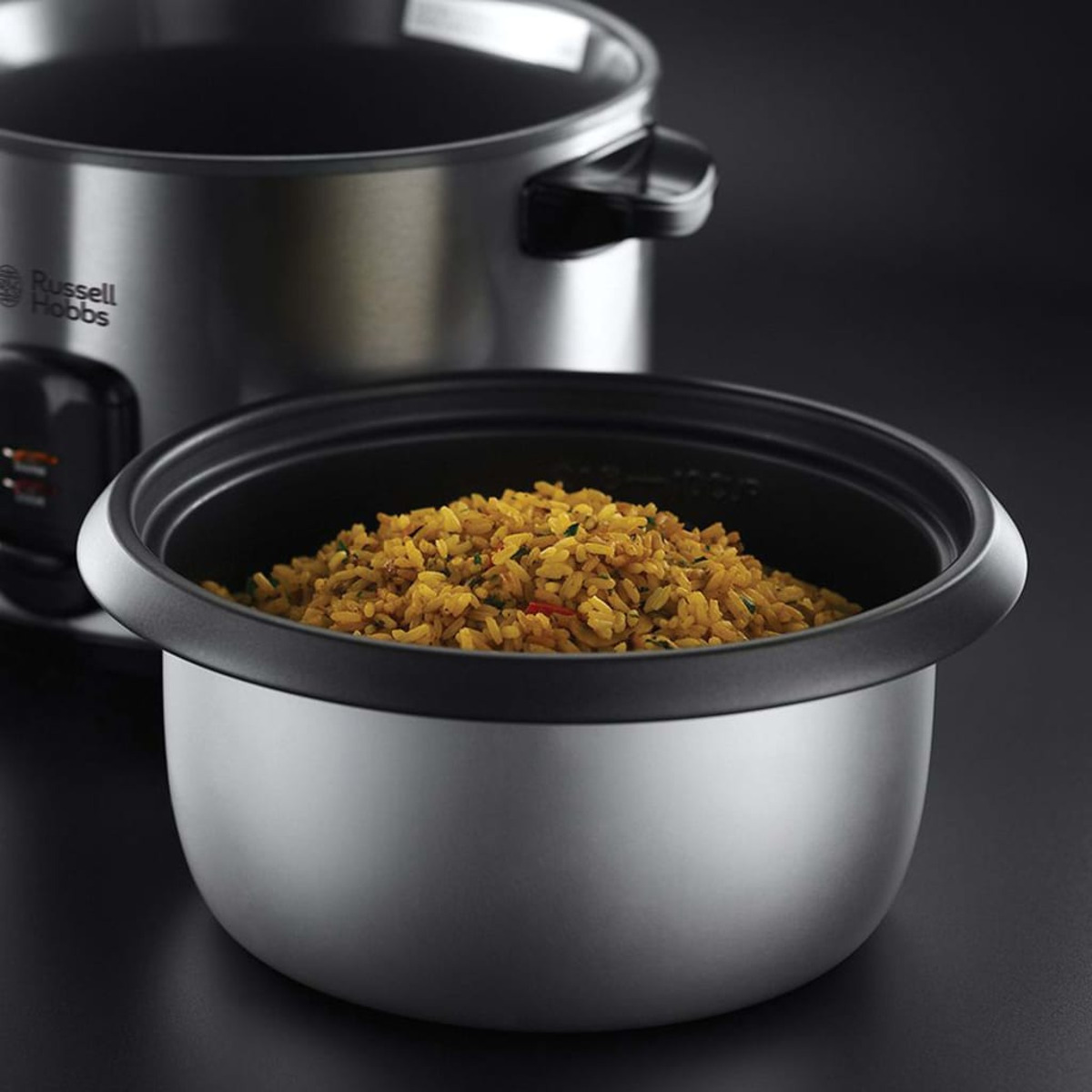 Urządzenie do gotowania ryżu z ugotowanym ryżem w środku, czarne tło, drugi garnek rozmyty w tle.