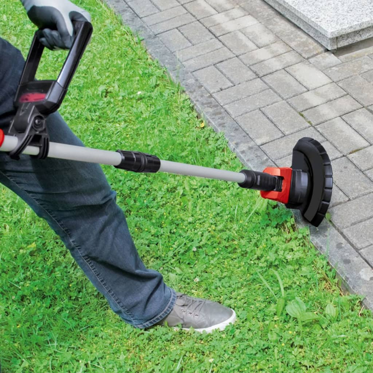 Person verwendet Rasentrimmer am gepflasterten Rand, grünes Gras und Betonhintergrund.