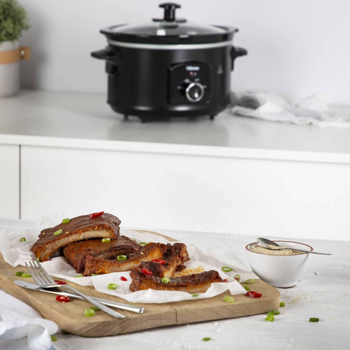 Una olla de cocción lenta negra con costillas cocidas, perejil y un cuenco de semillas de sésamo en una tabla de madera.