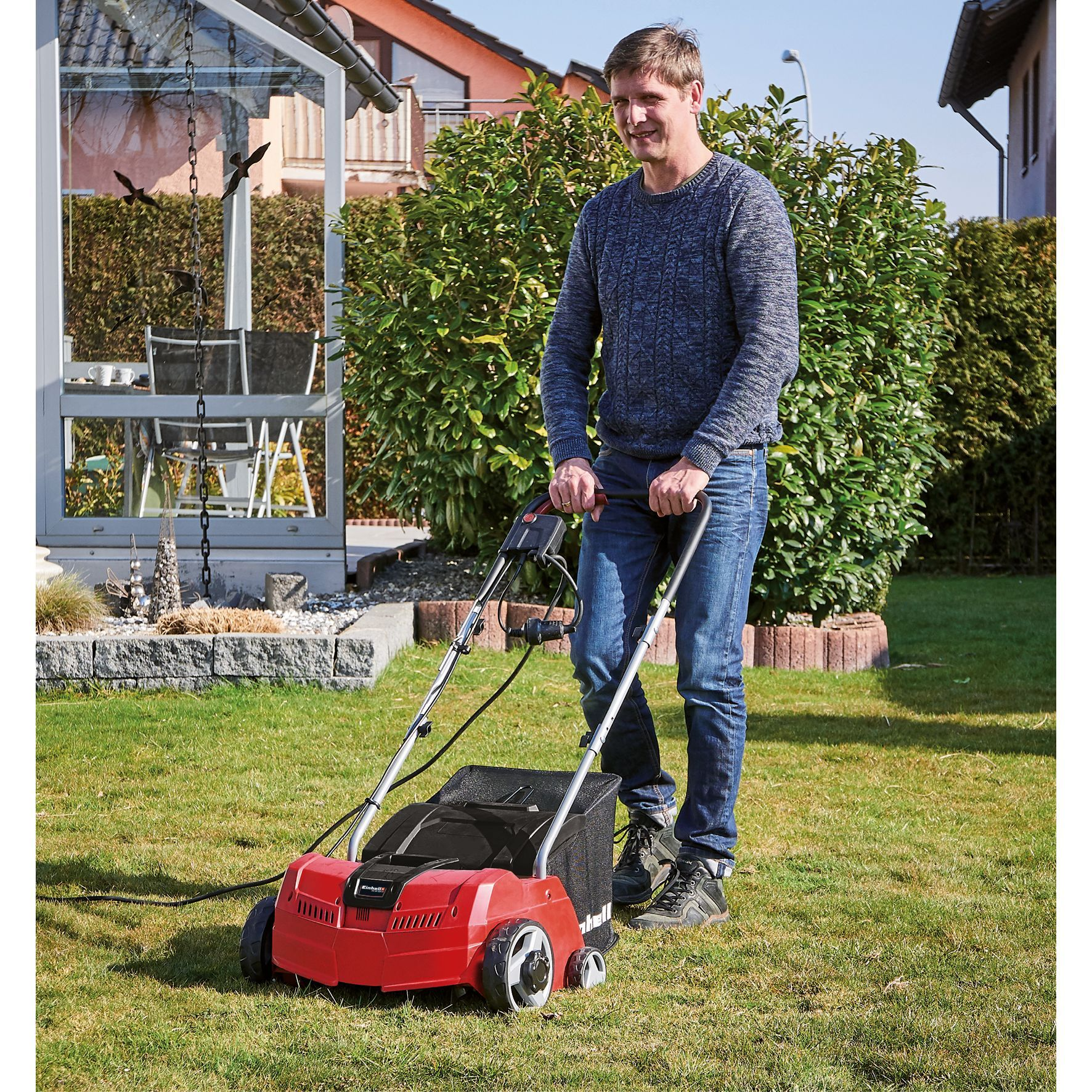 Ein Mann mäht einen Rasen mit einem roten Rasenlüfter. Er trägt Jeans und einen blauen Pullover.