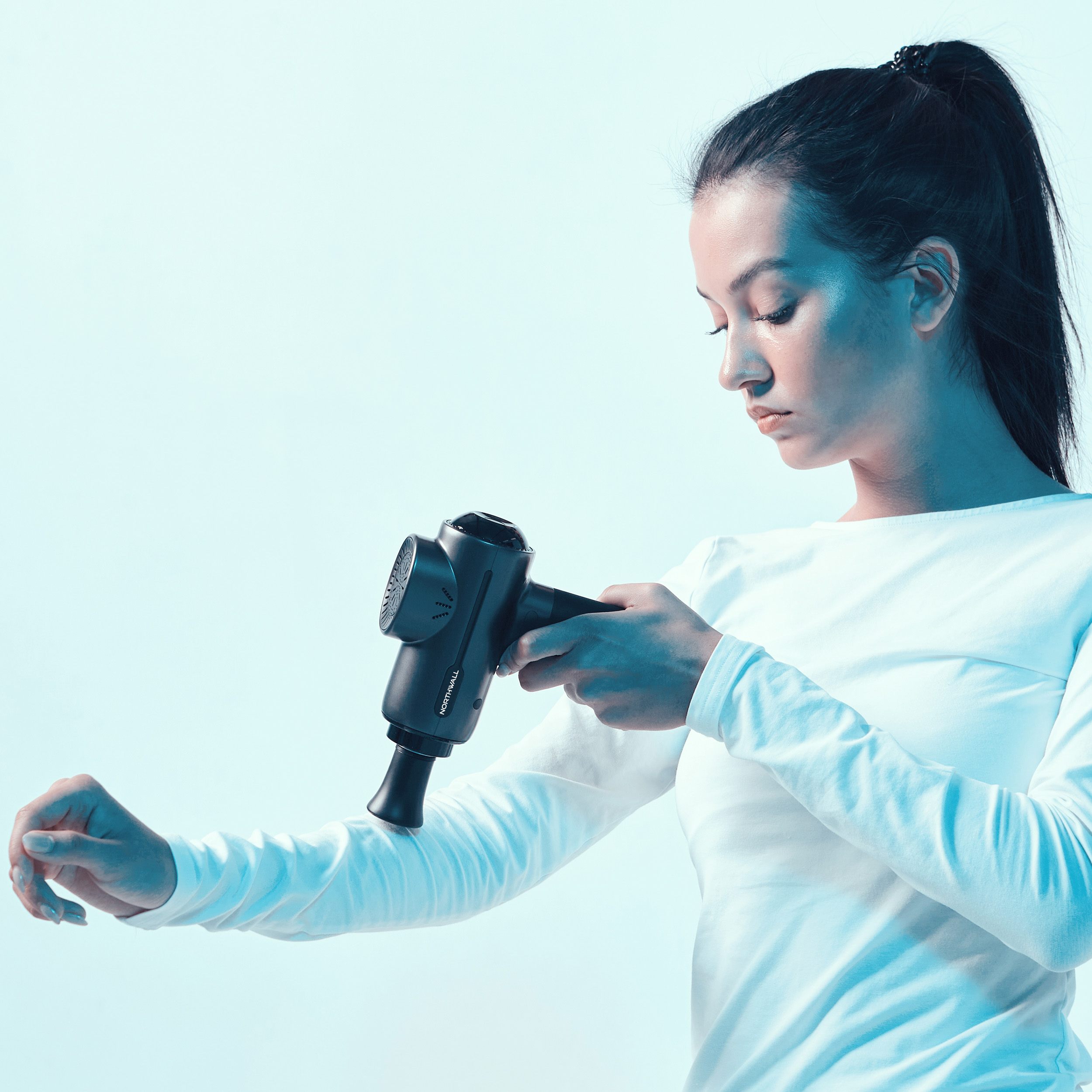 Frau, die eine schwarze Massagepistole an ihrem Arm vor hellblauem Hintergrund benutzt.