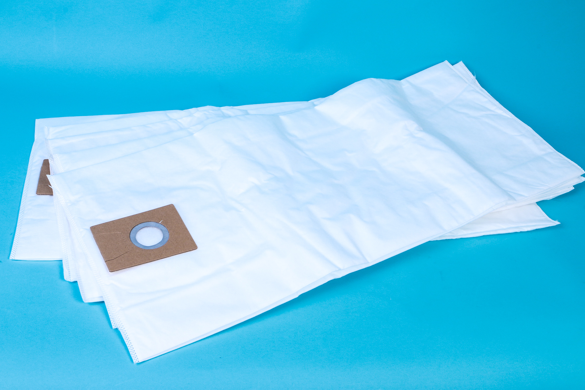 Weiße Staubsaugerbeutel mit braunem Kartonstück vor blauem Hintergrund.