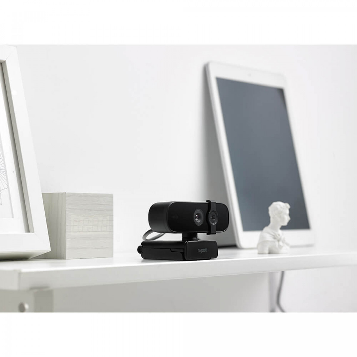 Czarna kamera internetowa na białej półce. Tablet i popiersie w tle.