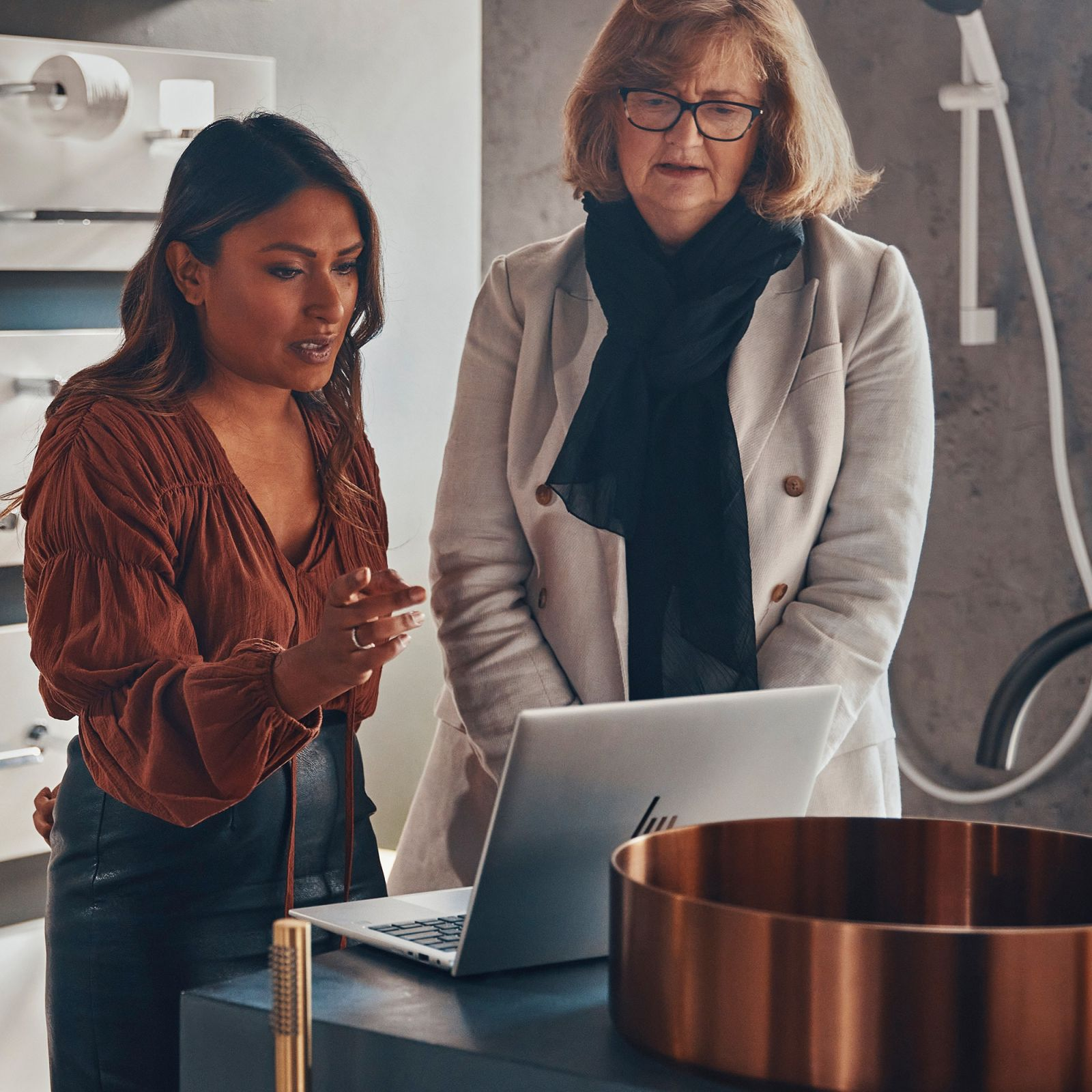 Dwie kobiety omawiają coś przed laptopem i miedzianą umywalką.