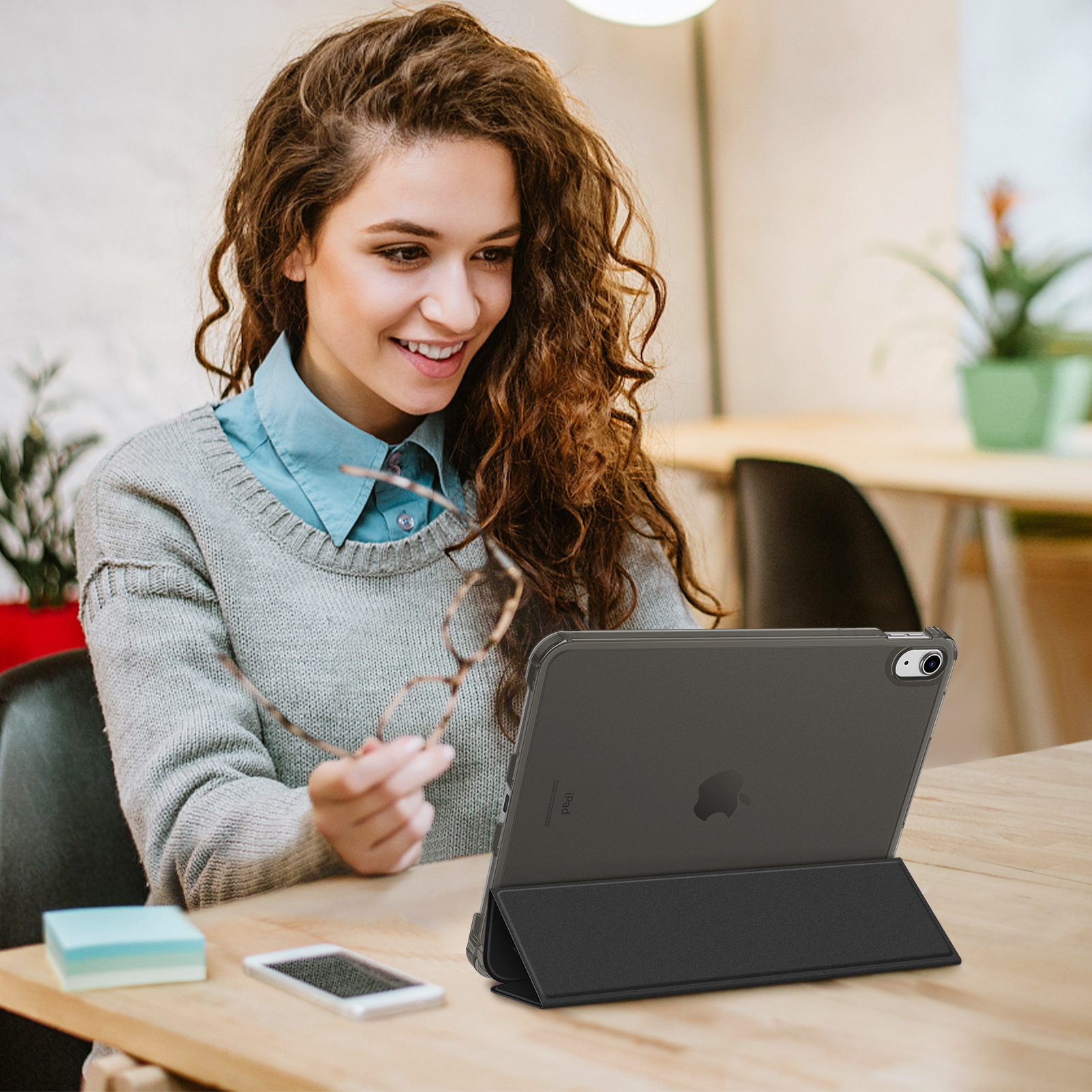 Frau benutzt Tablet mit Hülle am Schreibtisch. Lächelt, hält Brille. Bücher und Telefon sichtbar.