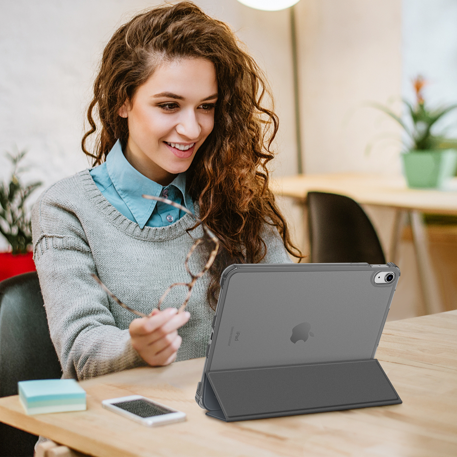 Eine Frau mit lockigem Haar betrachtet ein Tablet in einer Hülle, hält eine Brille, sitzt am Tisch.