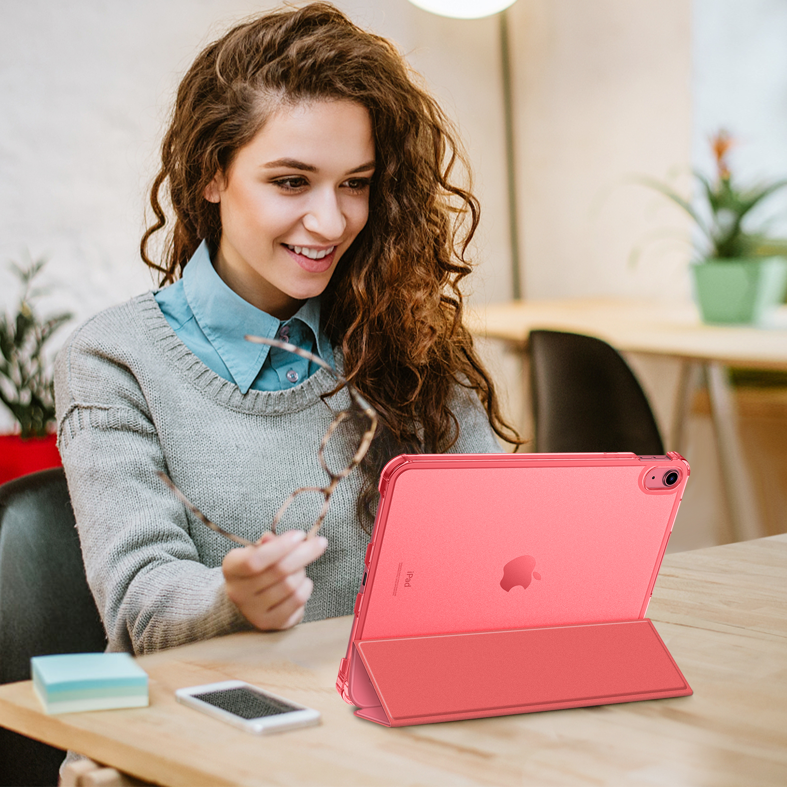 Eine Frau lächelt, während sie eine Brille hält, sie schaut auf ein rosa Tablet in einer Hülle auf einem Tisch.